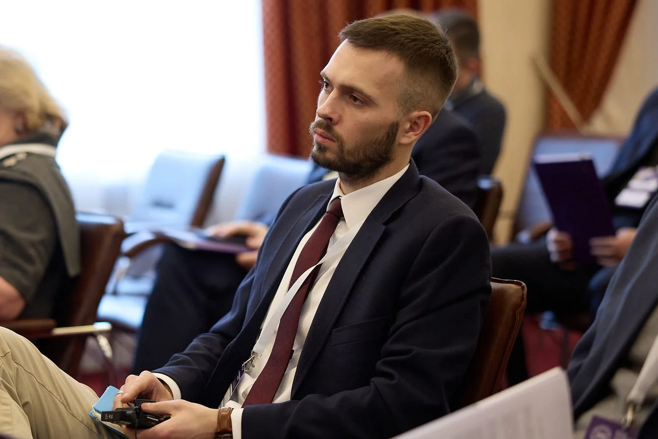 Młody mężczyzna z broda i wąsami, w ciemnej marynarce i bordowym krawacie.