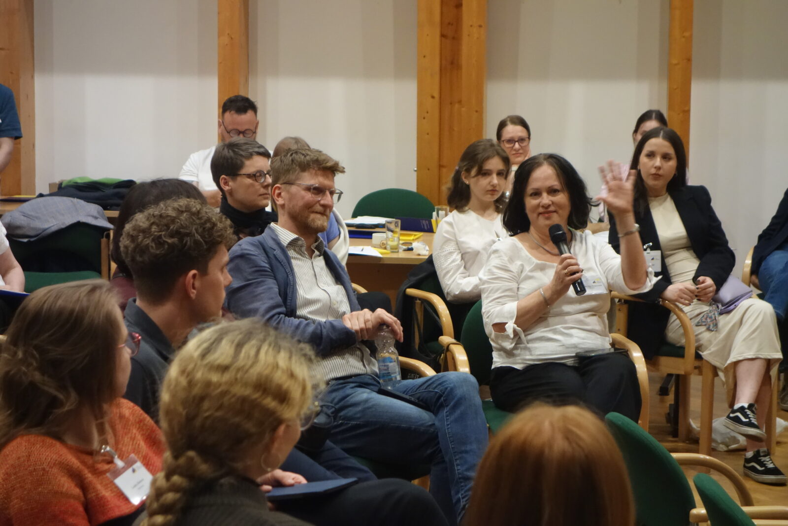 Grupa ludzi siedzi w sali konferencyjnej. Kobieta w średnim wieku trzyma w dłoni mikrofon.