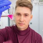 Młody mężczyzna trzymający w dłoni z założoną fioletową rękawiczką mały pojemnik z purpurową cieczą.