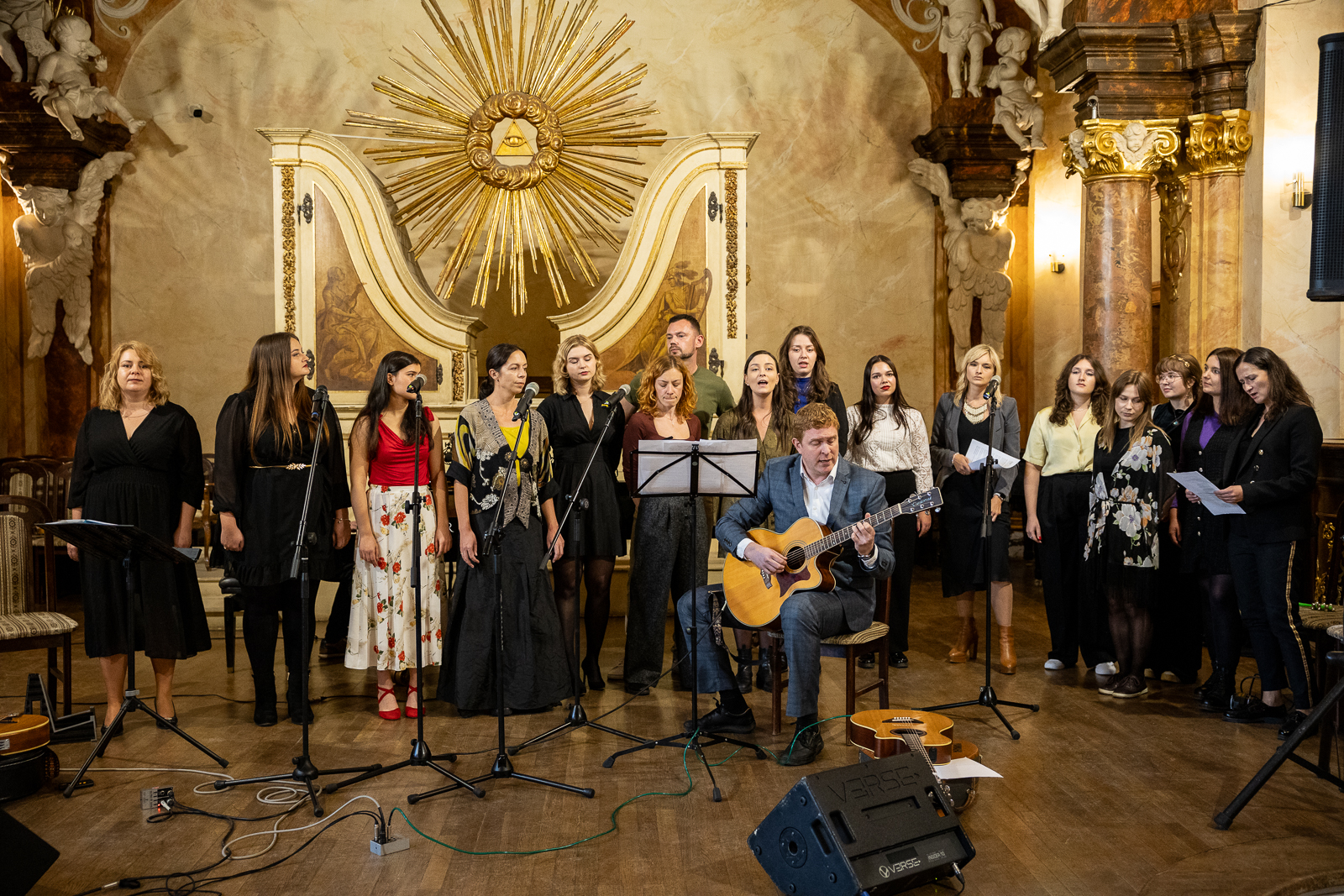 grupa ludzi stojąca przed mikrofonami w historycznym wnętrzu, n a środku mężczyzna z gitarą