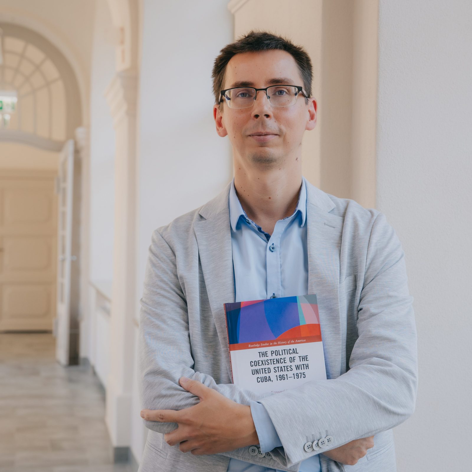 młody mężczyzna w jasnosiwej marynarce i niebieskiej koszuli, w okularach, trzyma w rekach książkę