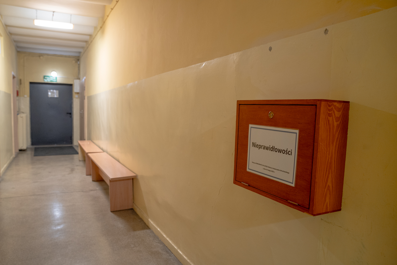 Korytarz oświetlony lampą, na żółtej ścianie wisi drewniana skrzynka z napisem "Nieprawidłowości"