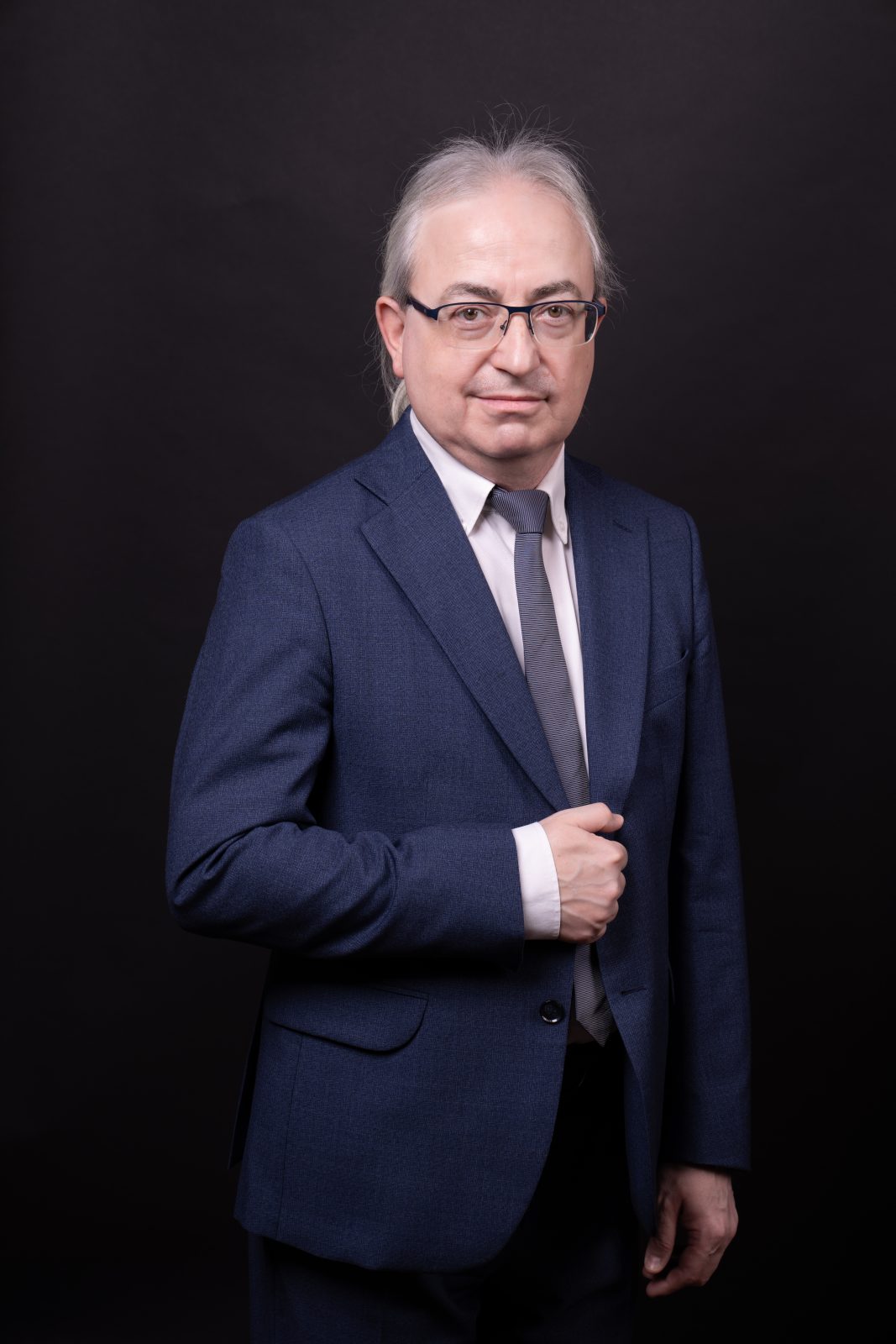 Mężczyzna z ręką na piersi, długimi i splecionymi w kucyk siwymi włosami i w okularach, ubrany w garnitur, stojący na czarnym tle. Dr hab. Filip Wolański, prof. UWr, członek Rady UWr na kadencję 2025-2028