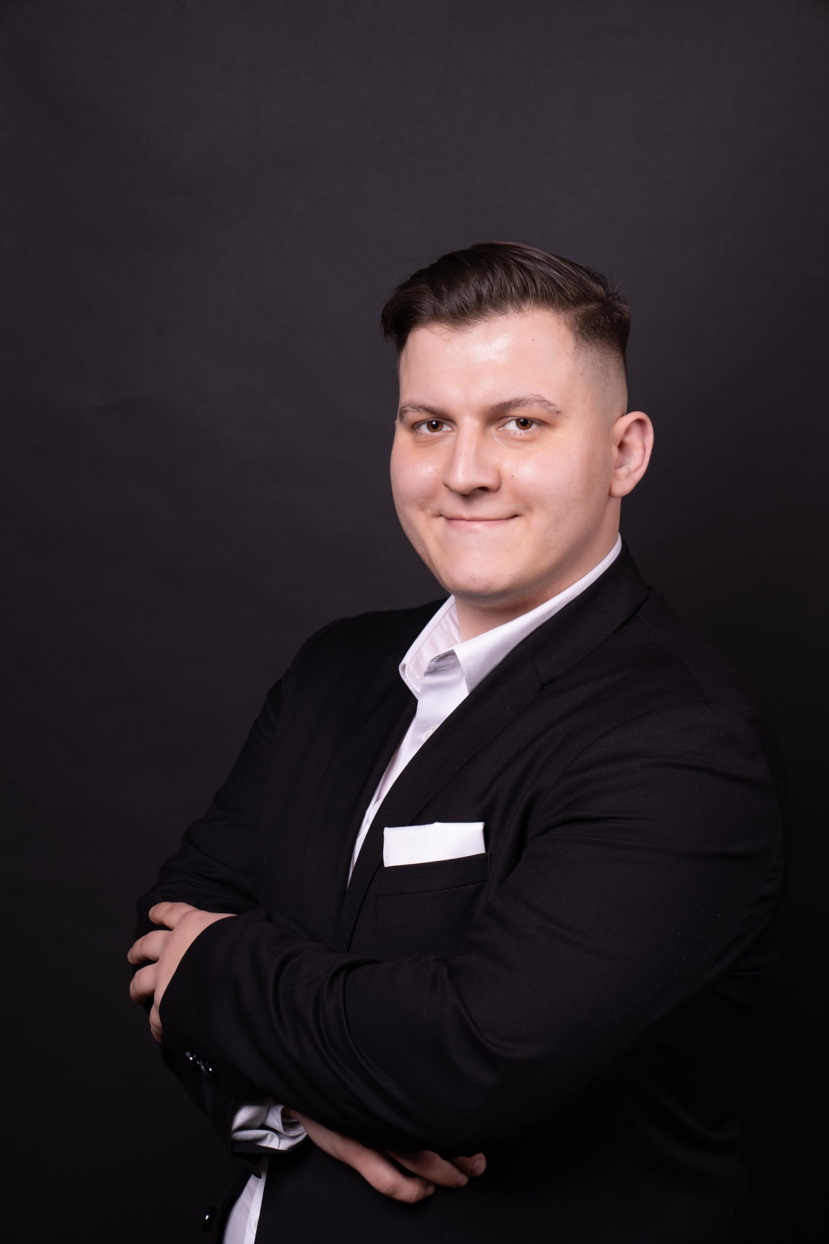 Młody mężczyzna ze splecionymi ramionami, ubrany w garnitur, stojący na czarnym tle. Arkadiusz Smugowski, przewodniczący Samorządu Studentów UWr, członek Rady UWr na kadencję 2025-2028
