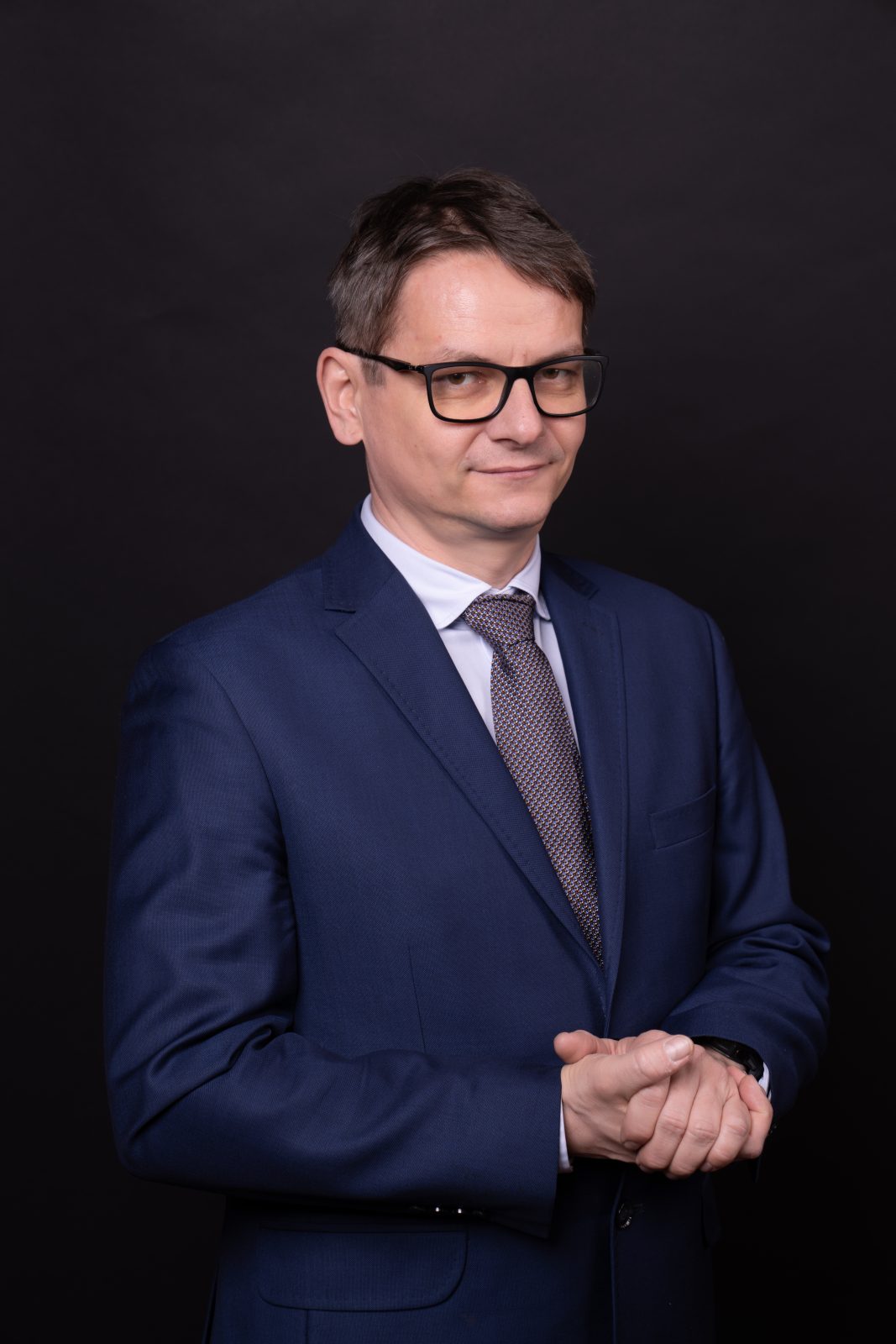 Mężczyzna ze splecionymi dłońmi, ubrany w garnitur, stojący na czarnym tle. Dr hab. Krzysztof Graczyk, prof. UWr, członek Rady UWr na kadencję 2025-2028