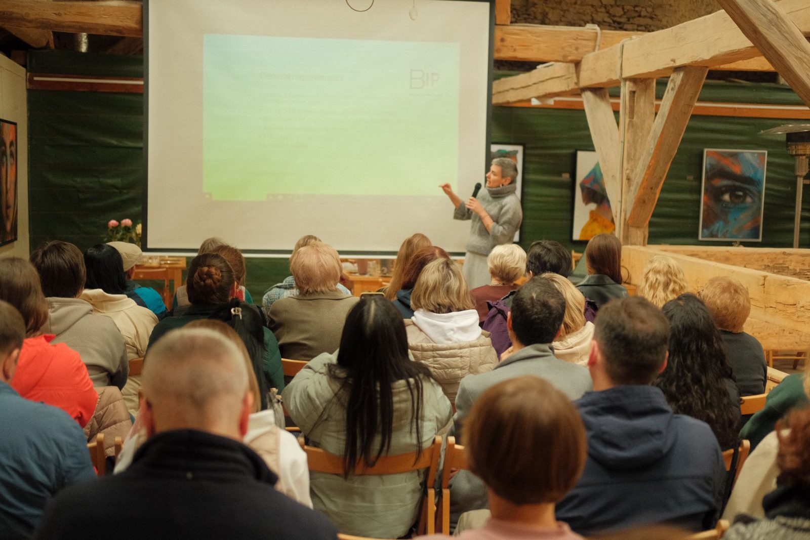 Sala z ludźmi siedzącymi na krzesłach widocznymi od tyłu, w głębi sali kobieta w średnim wieku wskazuje ręką na biały ekran do projektora z wyświetlonym niewyraźnym tekstem