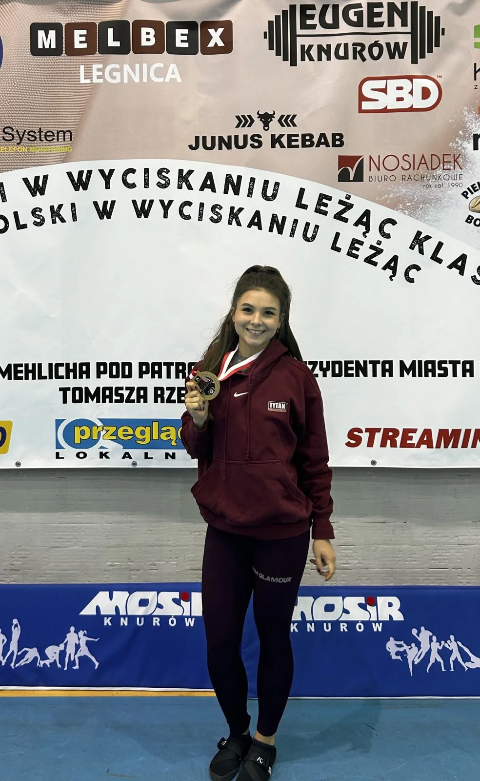 Młoda kobieta z długimi włosami stoi trzymając w dłoni zawieszony na szyi złoty medal