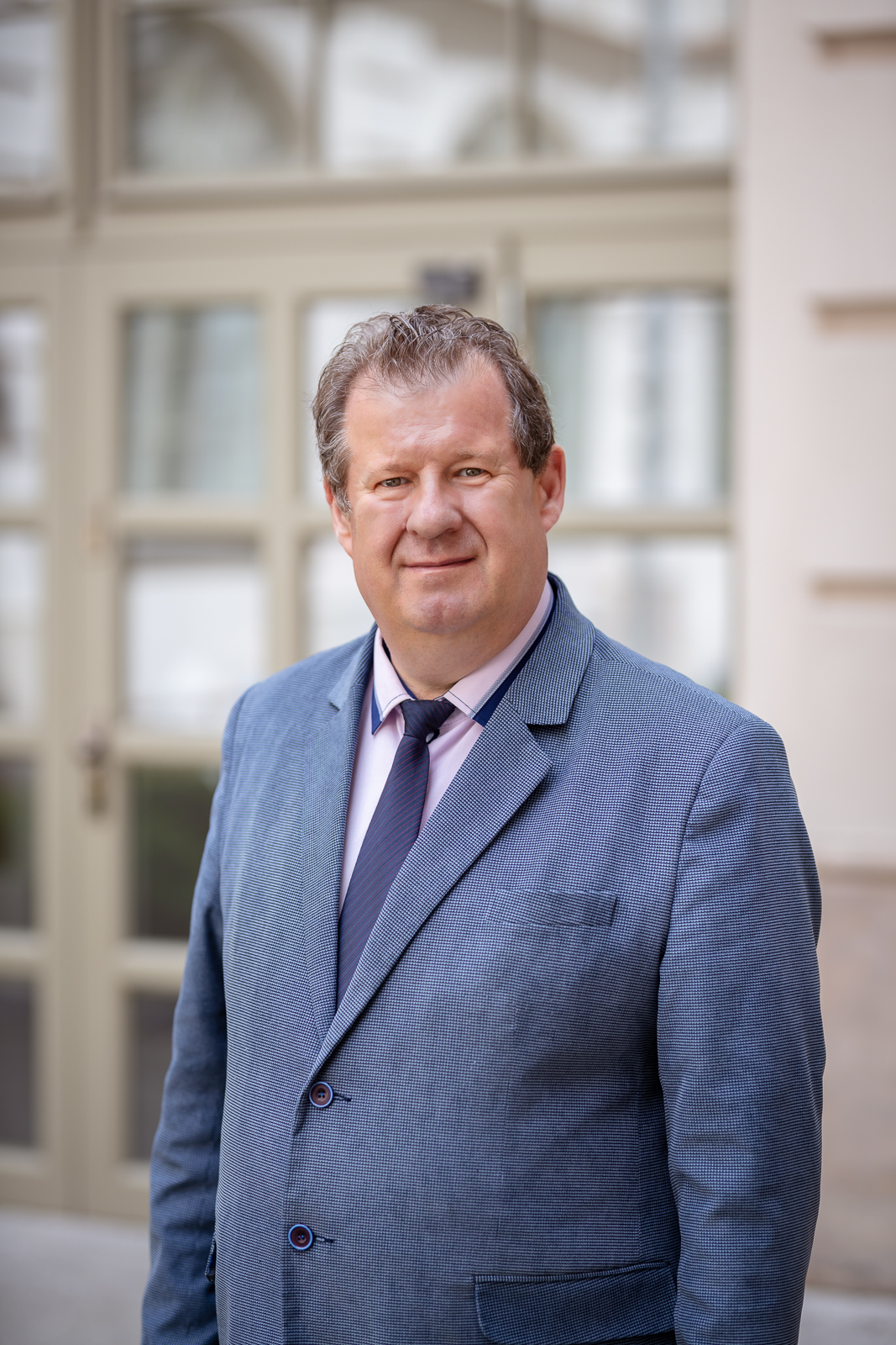 Mężczyzna z lekko kręconymi włosami, w niebieskim garniturze, białej koszuli i granatowym krawacie.
