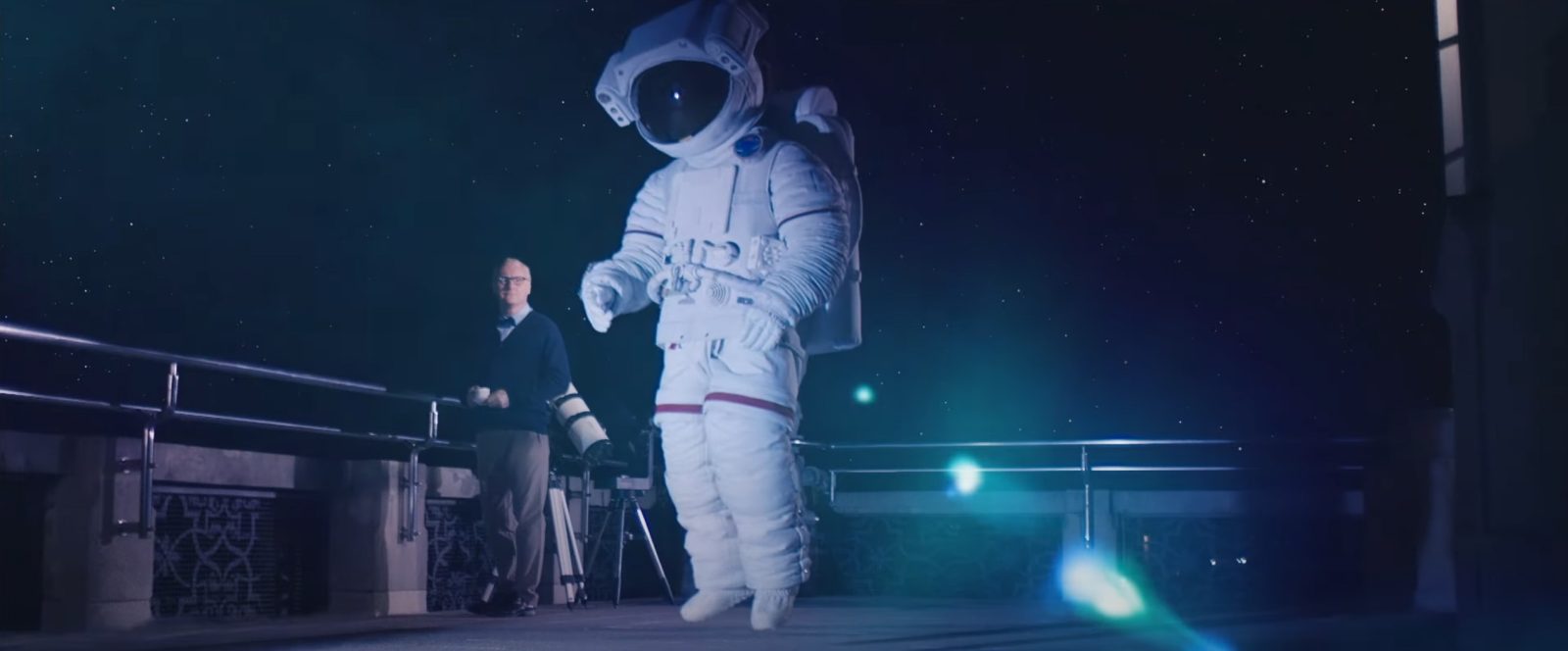 astronauta w ciemności, po lewej stronie, z tyłu, stoi siwy mężczyzna w okularach, za nm barierka