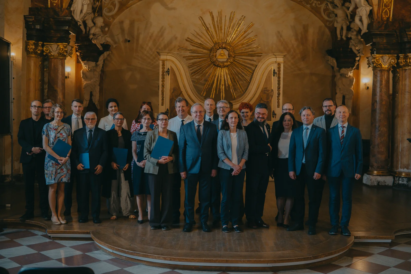 Grupa dwudziestu kilku osób w średnim wieku, stojących na scenie w sali ze złotymi Zdobieniami na ścianach