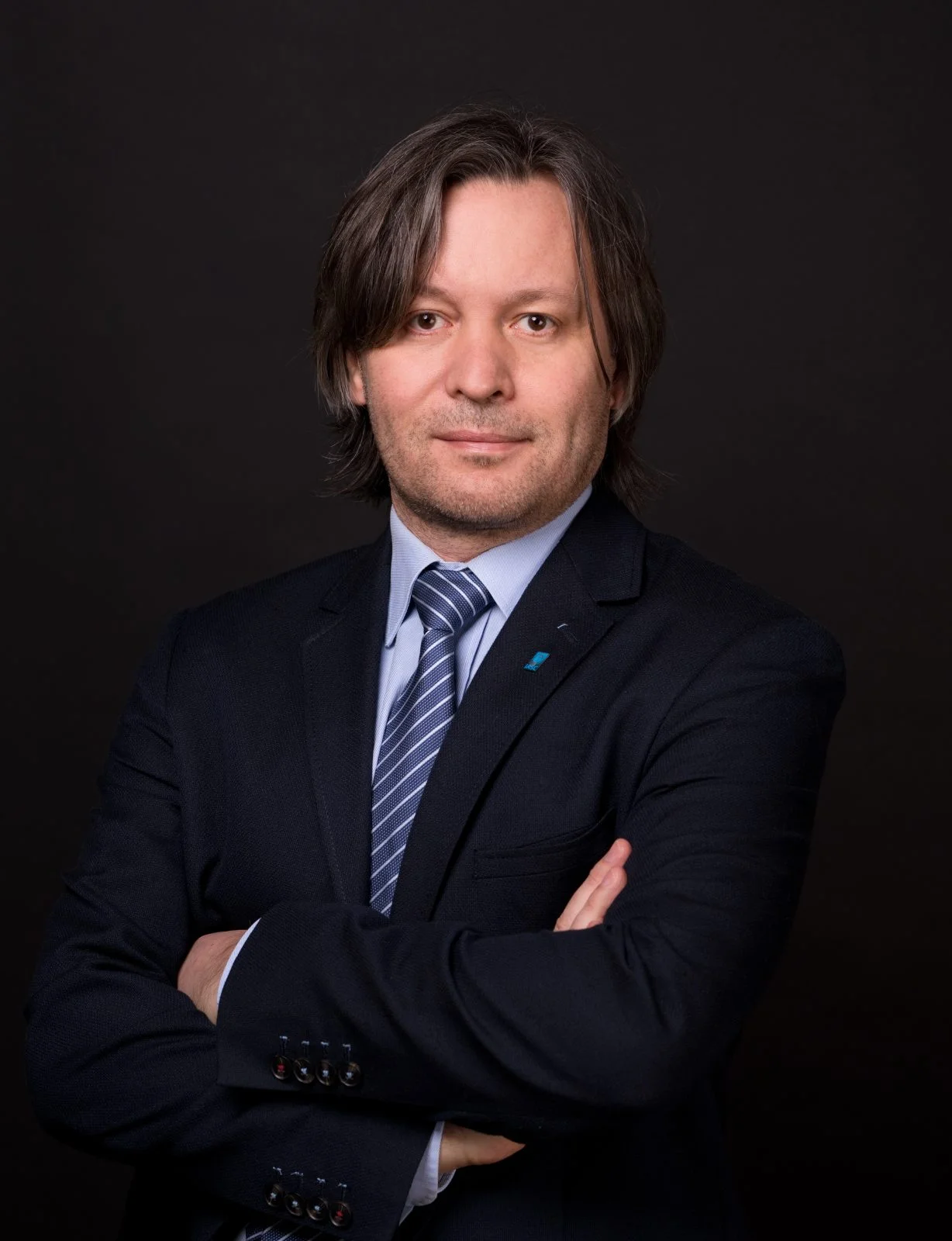 Mężczyzna z dłuższymi włosami w ciemnym garniturze, niebieskim krawacie i jasnoniebieskiej koszuli stoi na ciemnym tle z założonymi rękami.