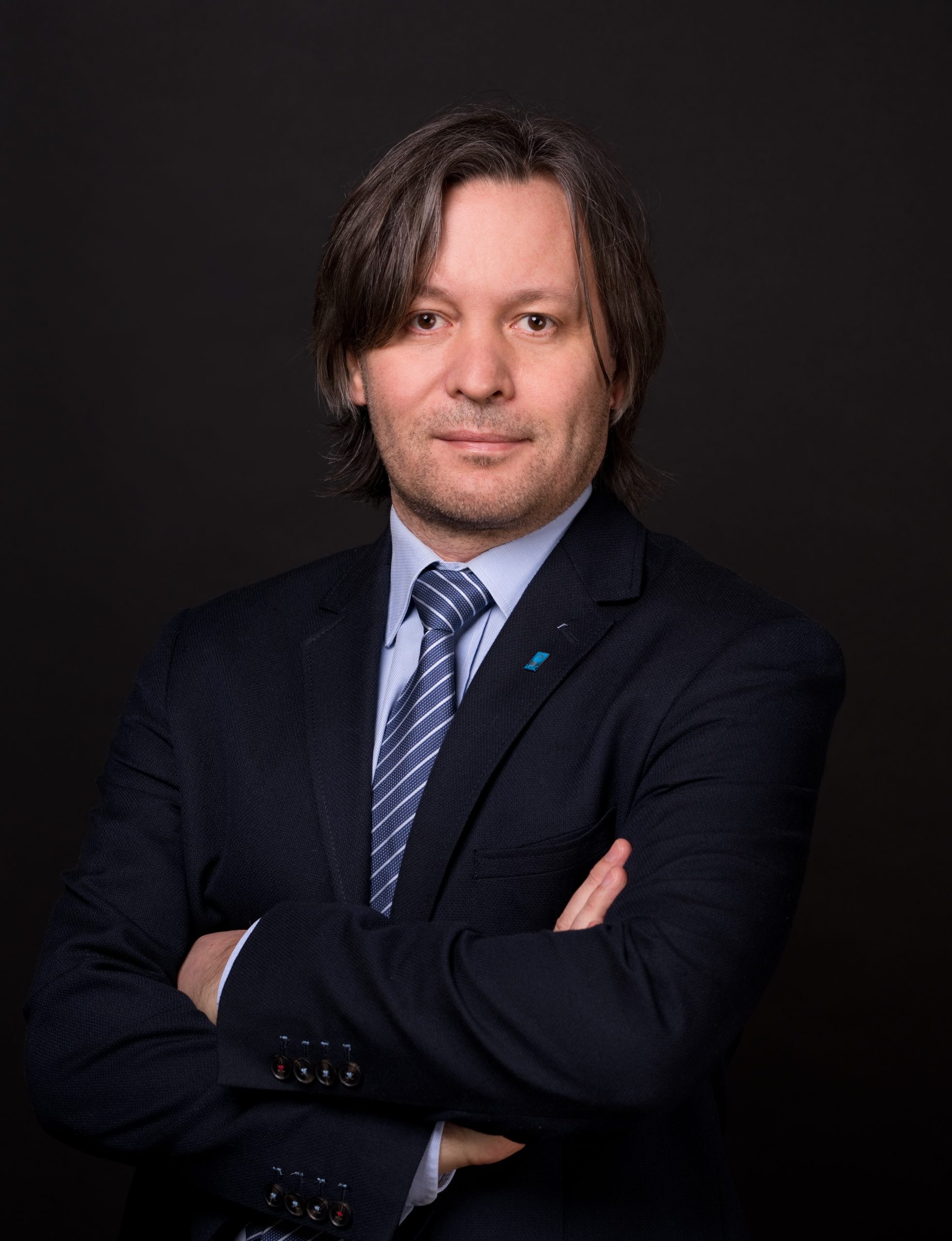 Mężczyzna z dłuższymi włosami w ciemnym garniturze, niebieskim krawacie i jasnoniebieskiej koszuli stoi na ciemnym tle z założonymi rękami.