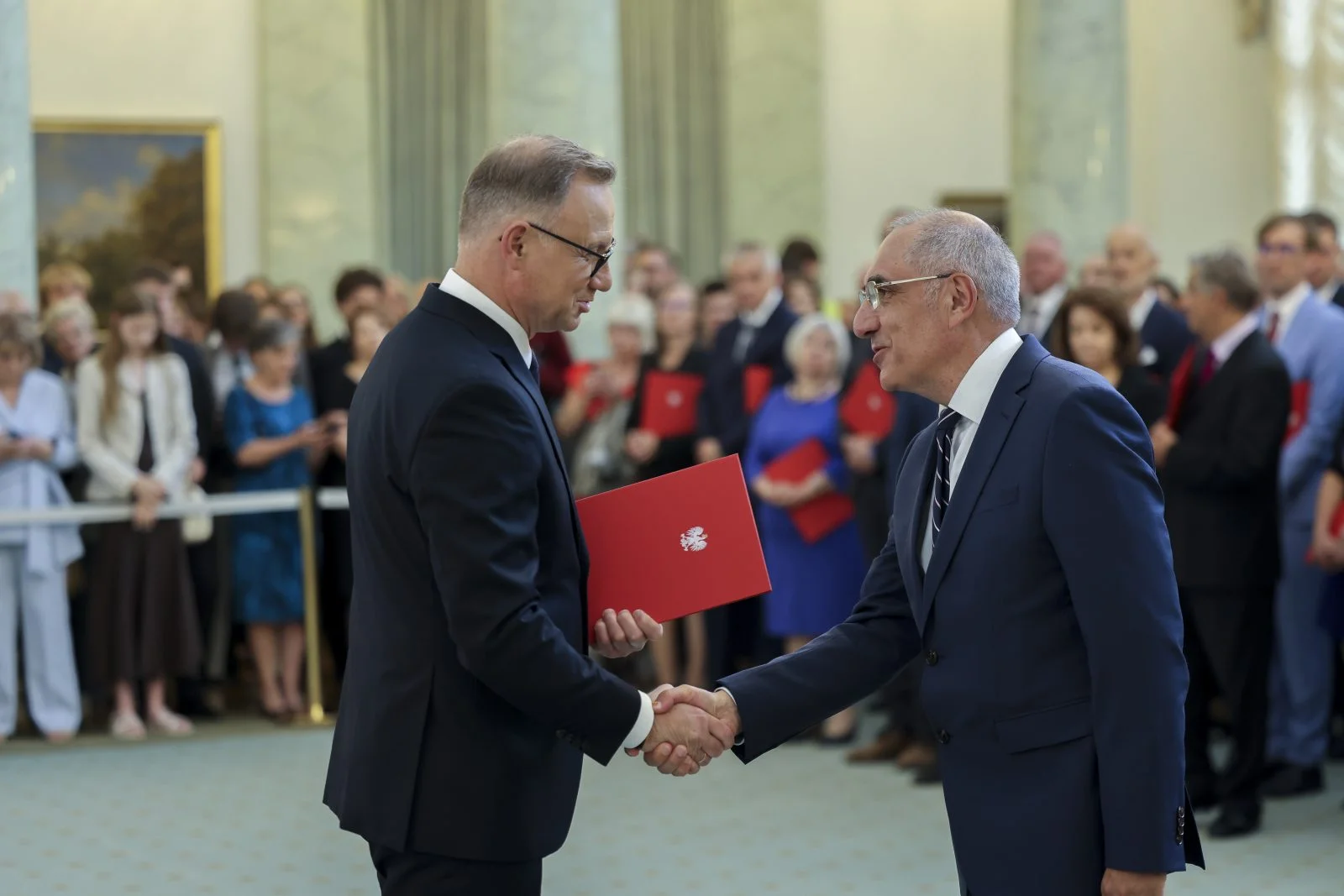 dwóch mężczyzn ubranych wizytowo, jeden wręcza drugiemu nagrodę w pałacu prezydenckim