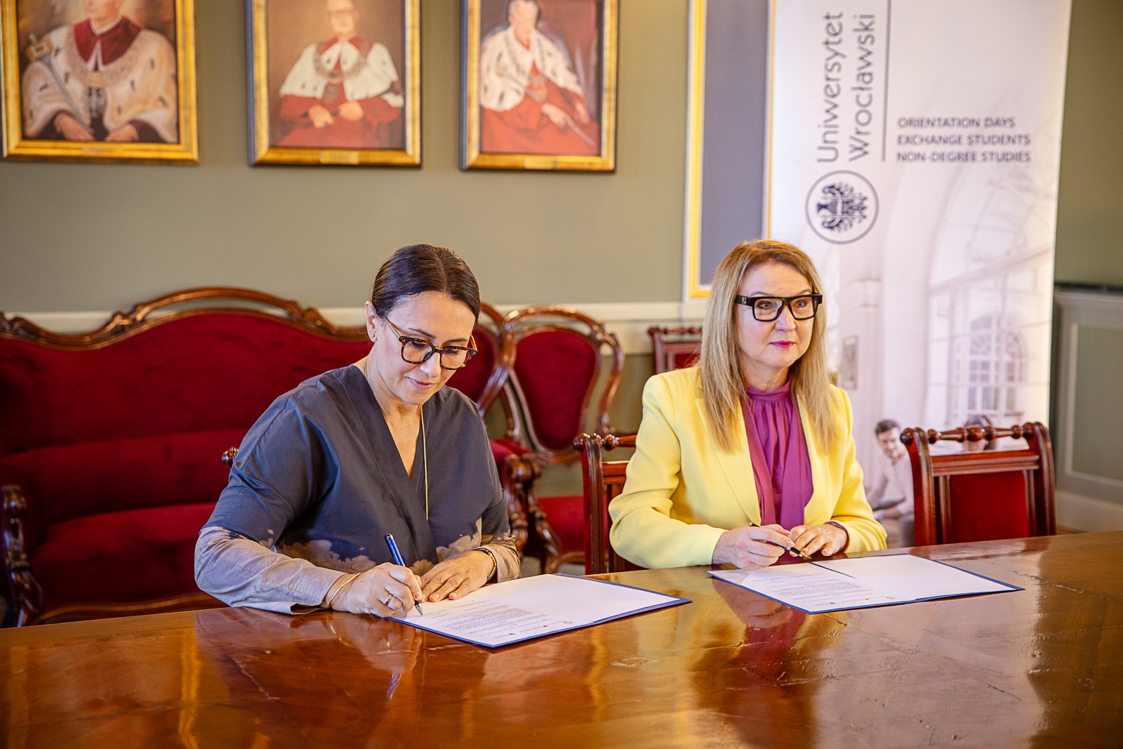 Dwie kobiety w średnim wieku siedzą przy dużym drewnianym stole, przed nimi dokumenty
