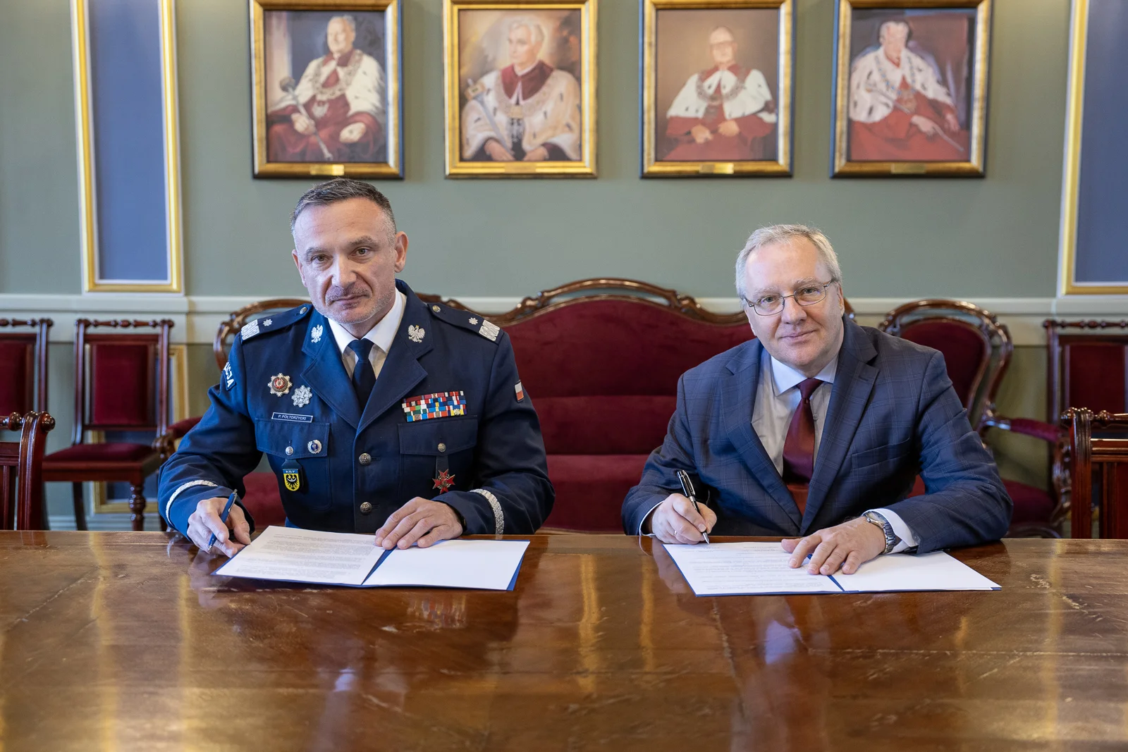 Dwóch mężczyzn w średnim wieku siedzi przy stole i podpisuje dokumenty. Jeden z nich ubrany jest w uroczysty, policyjny mundur, drugi w grafitowy garnitur. W tle widać ozdobne meble i obrazy przedstawiające byłych rektorów uniwersytetu.