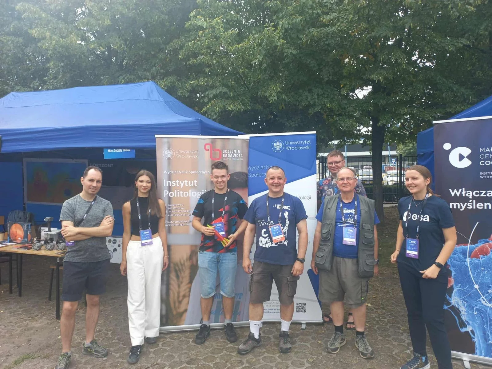 Na zdjęciu widać grupę siedmiu osób stojących przed niebieskimi namiotami wystawowymi. Za nimi ustawione są banery Uniwersytetu Wrocławskiego, Instytutu Politologii i Europejskiej Agencji Kosmicznej. Część osób nosi koszulki z logotypem ESA, inni ubrani są bardziej swobodnie. W tle na stoliku widać modele naukowe, w tym miniaturowego łazika. Cała grupa uśmiecha się do kamery.