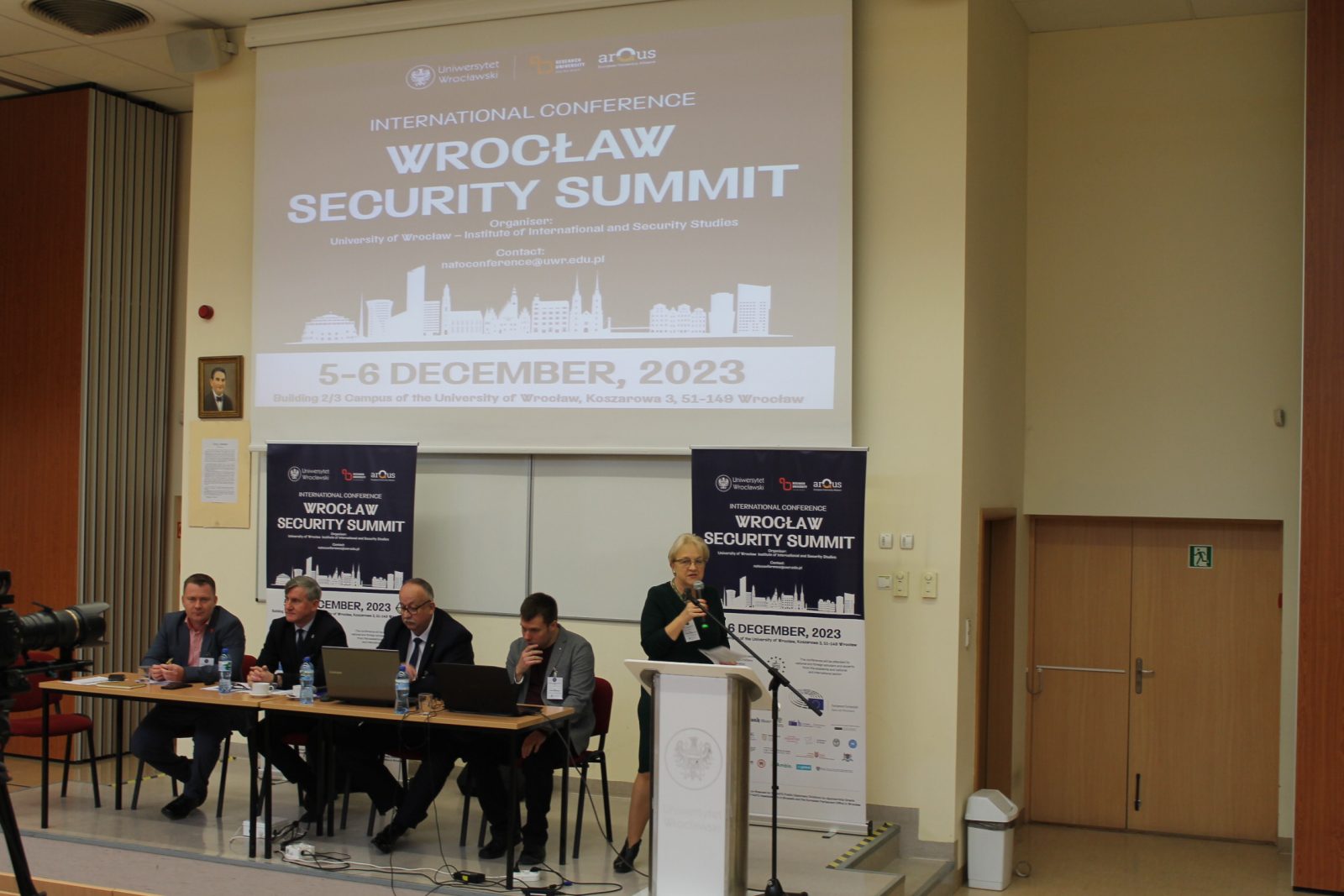 Na zdjęciu widać scenę z konferencji Wrocław Security Summit. Przy stole siedzi czterech prelegentów, a kobieta stojąca przy mównicy przemawia do publiczności; w tle widoczny jest ekran z informacjami o wydarzeniu oraz banery konferencyjne. Całość odbywa się w nowoczesnej sali wykładowej, z kamerą nagrywającą wydarzenie.
