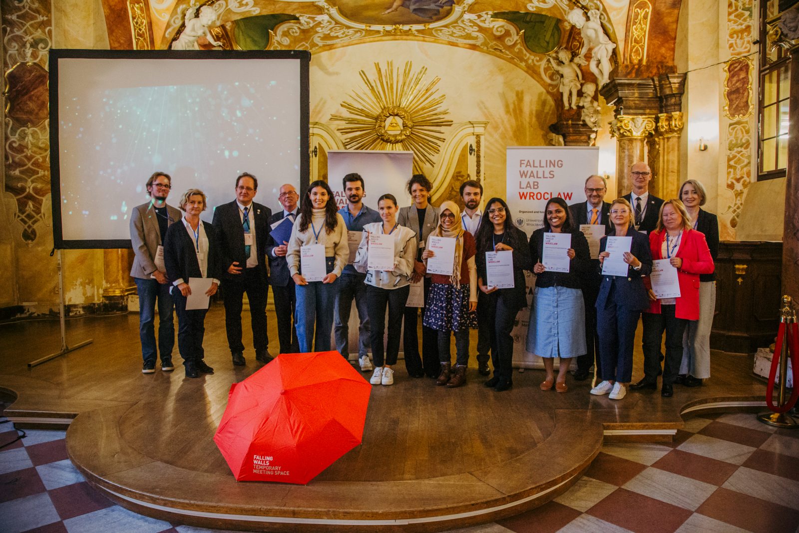 grupa ludzi stoi w historycznej sali, przed nimi czerwony parasol, 