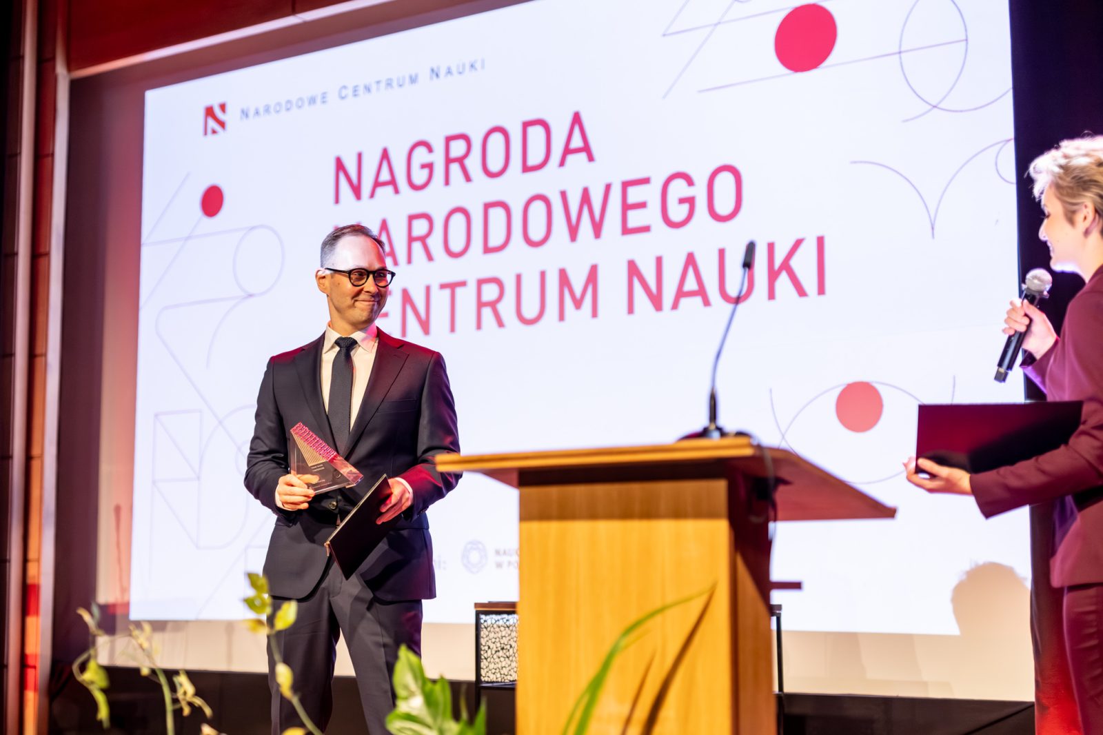 mężczyzna w średnim wieku w sali konferencyjnej z ekranem z napisem Nagroda narodowego centrum nauki
