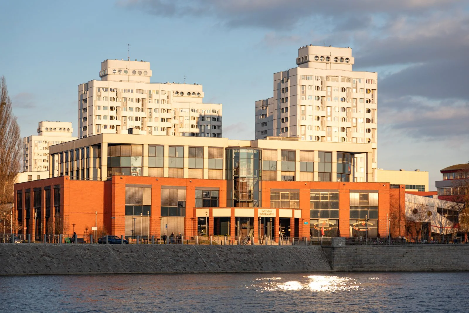Nowoczesny budynek z czerwonej cegły i dużych przeszklonych powierzchni. Na jego froncie widnieje napis ‘Instytut Informatyki’. Za nim wznoszą się wysokie, jasne bloki mieszkalne, tzw. sedesowce.
