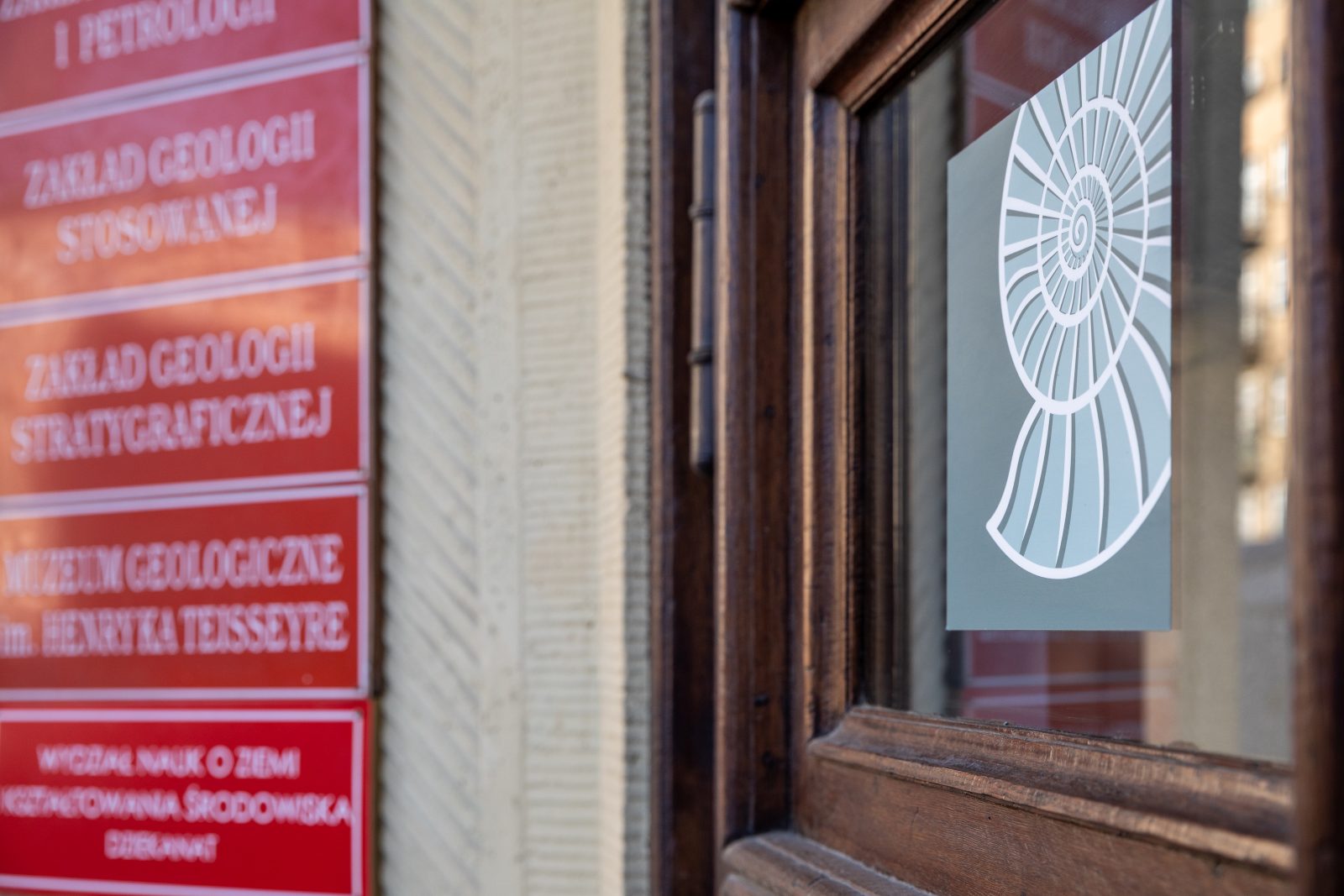 Drzwi z szybą i naklejoną na niej grafiką amonitu. Obok czerwone tabliczki z rozmazanymi nazwami instytucji.