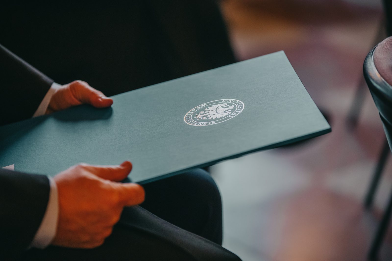 Zdjęcie przedstawia elegancką, zieloną teczkę z białym emblematem Uniwersytetu Wrocławskiego na okładce, trzymaną przez dłonie osoby siedzącej na krześle.