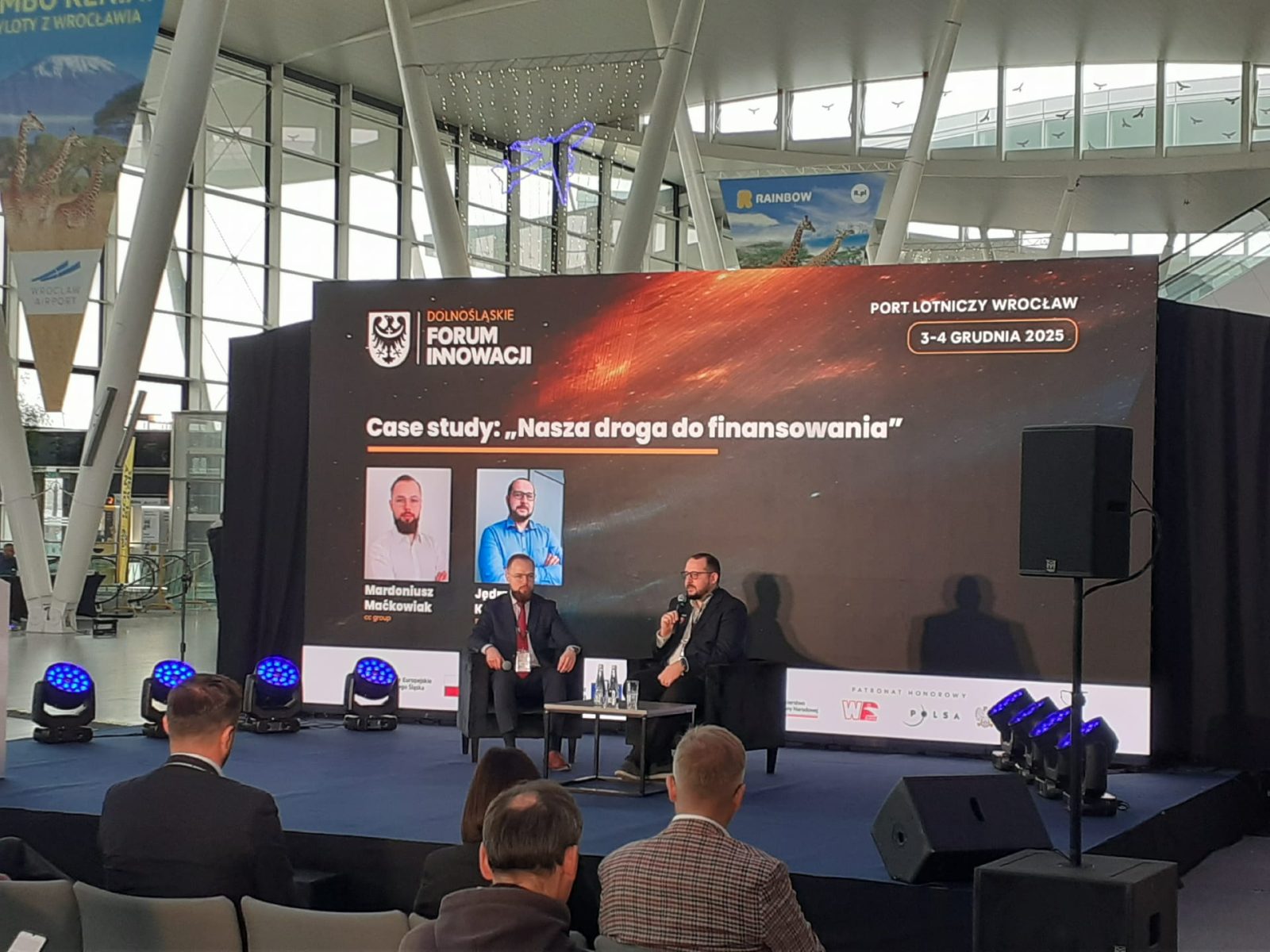 Zdjęcie przedstawia scenę wydarzenia Dolnośląskie Forum Innowacji, które odbywa się w przestrzeni Portu Lotniczego Wrocław. Na scenie ustawione są dwa fotele i stolik, na których siedzą dwie osoby prowadzące rozmowę w formie case study zatytułowanej „Nasza droga do finansowania”. W tle znajduje się duży ekran z grafiką w odcieniach czerni, pomarańczu i czerwieni, na którym widnieją logo wydarzenia: „Dolnośląskie Forum Innowacji”, nazwa miejsca i daty: „Port Lotniczy Wrocław, 3–4 grudnia 2025” oraz tytuł sesji: „Case study: Nasza droga do finansowania”. Przed sceną siedzi kilku uczestników, a w tle widać nowoczesną, przeszkloną architekturę terminalu lotniczego z banerami reklamowymi i dekoracjami.