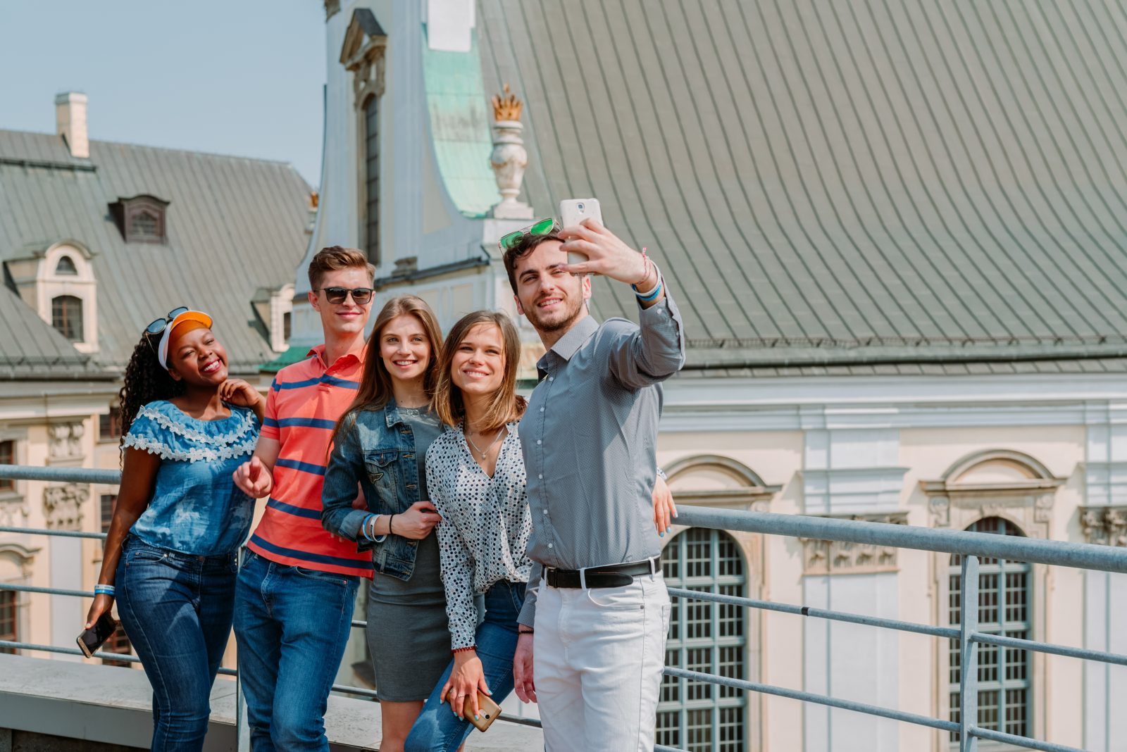 Grupa pięciu osób stoi na zewnątrz, pozując do wspólnego selfie. Jedna z osób trzyma telefon wyciągnięty przed siebie, a pozostali stoją blisko siebie, uśmiechając się i obejmując nawzajem. W tle widoczne są jasne, zabytkowe budynki z dużym, skośnym dachem i ozdobnymi fasadami.
