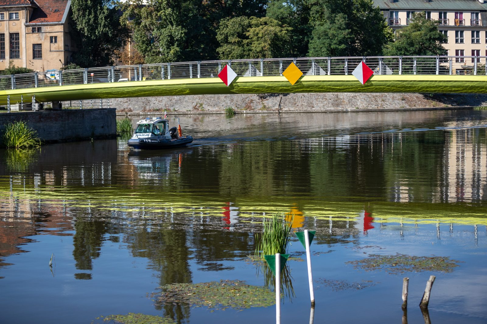 fragment rzeki z widocznym mostem pieszym w tle, z metalową balustradą i oznaczeniami nawigacyjnymi w postaci dużych tablic w kształcie rombów w kolorach czerwonym, białym i żółtym. Pod mostem płynie niewielka łódź policyjna z napisem „Policja” na burcie, wyposażona w pomarańczową flagę i sprzęt ratunkowy