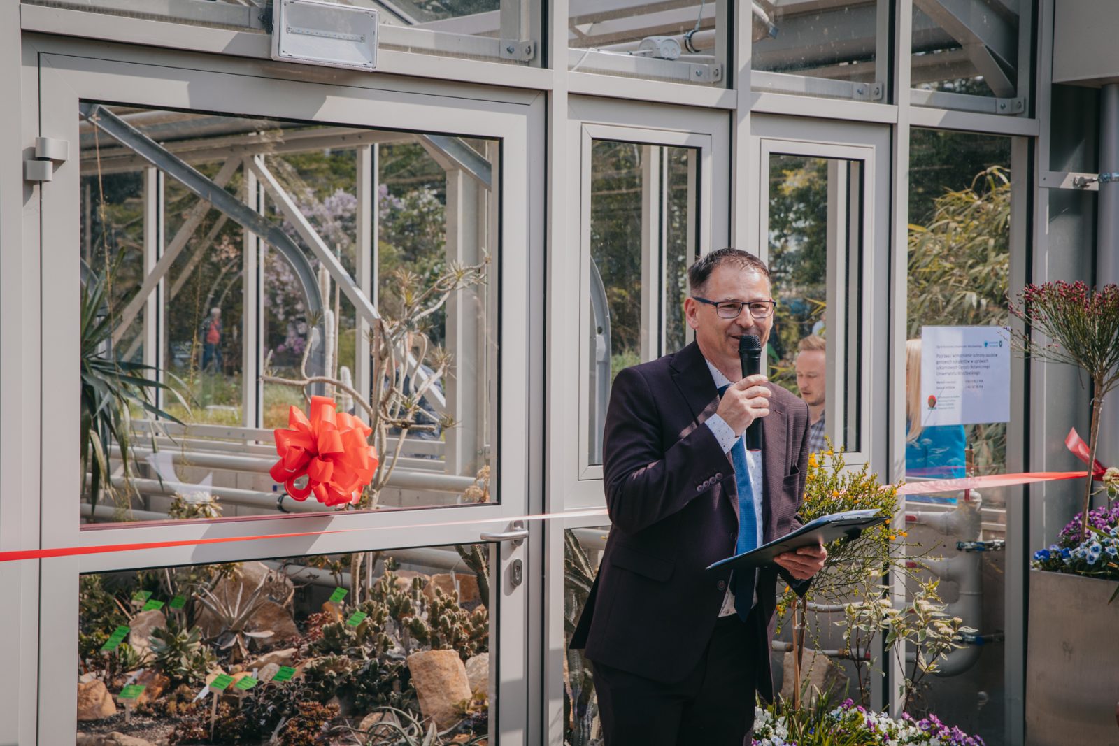 zdjęcie przedstawiające dojrzałego mężczyznę ubranego wizytowo, stoi przed szklarnią w ogrodzie