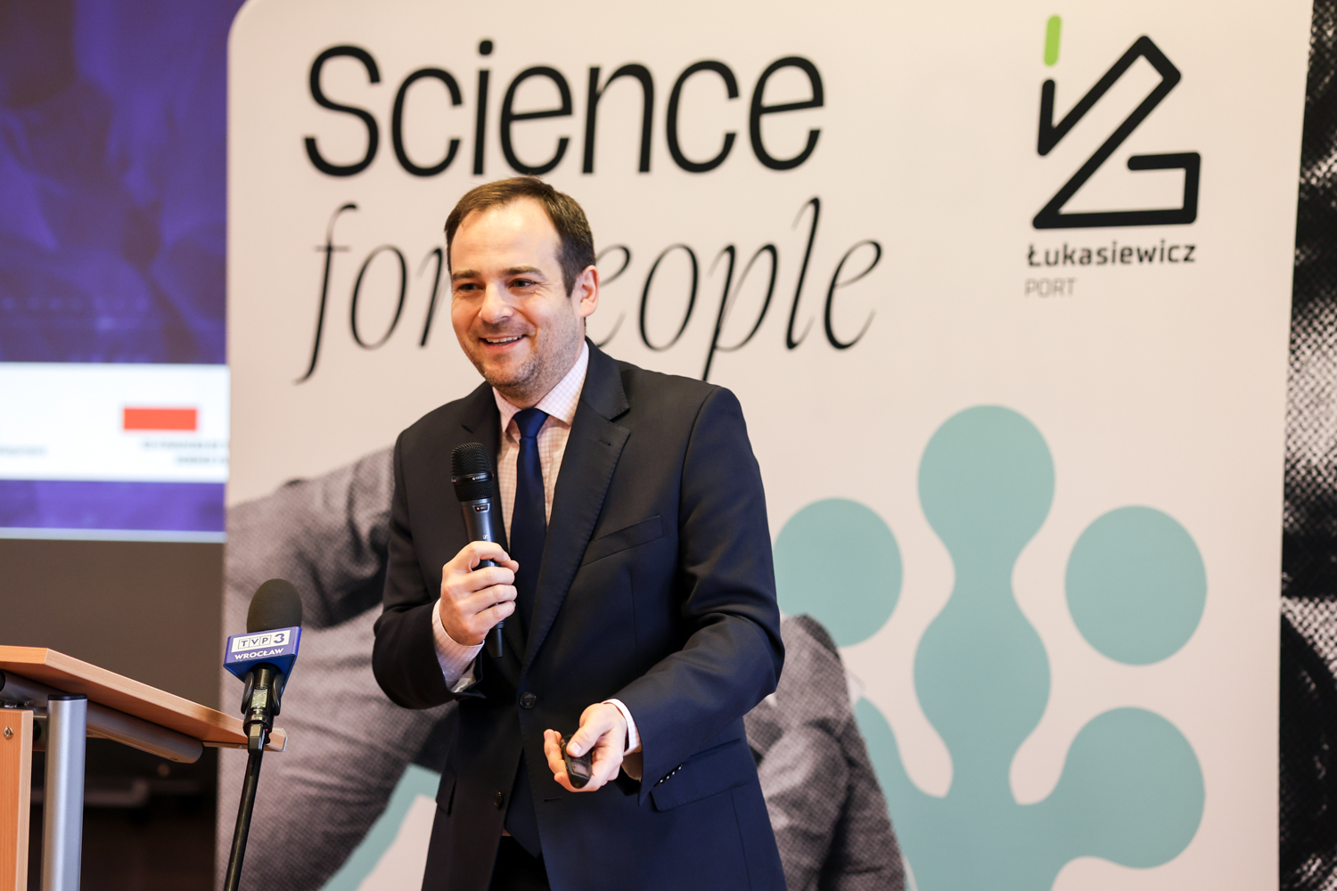 Mężczyzna ubrany w garnitur przemawiający do widowni za pomocą mikrofonu