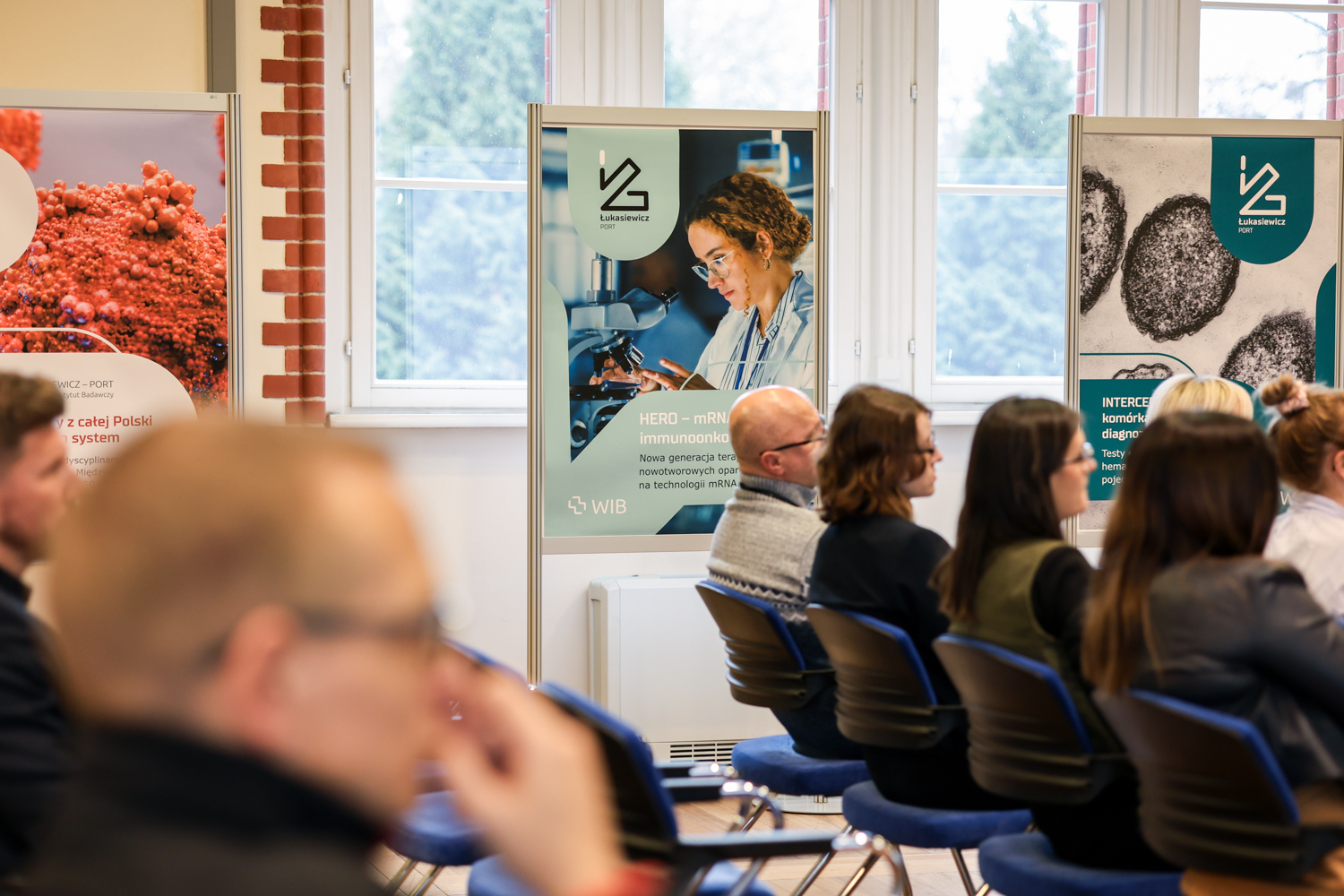 Grupa osób siedząca na krzesłach, odwrócona bokiem. Za nimi widoczne są trzy plakaty na dużych stojakach.