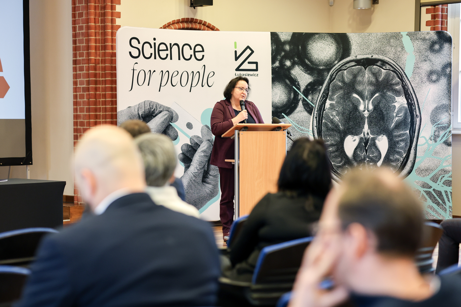 Kobieta przemawiająca do mikrofonu z mównicy, stojąca na tle dużej ścianki do zdjęć z napisem Science for People i fotografią ludzkiego mózgu