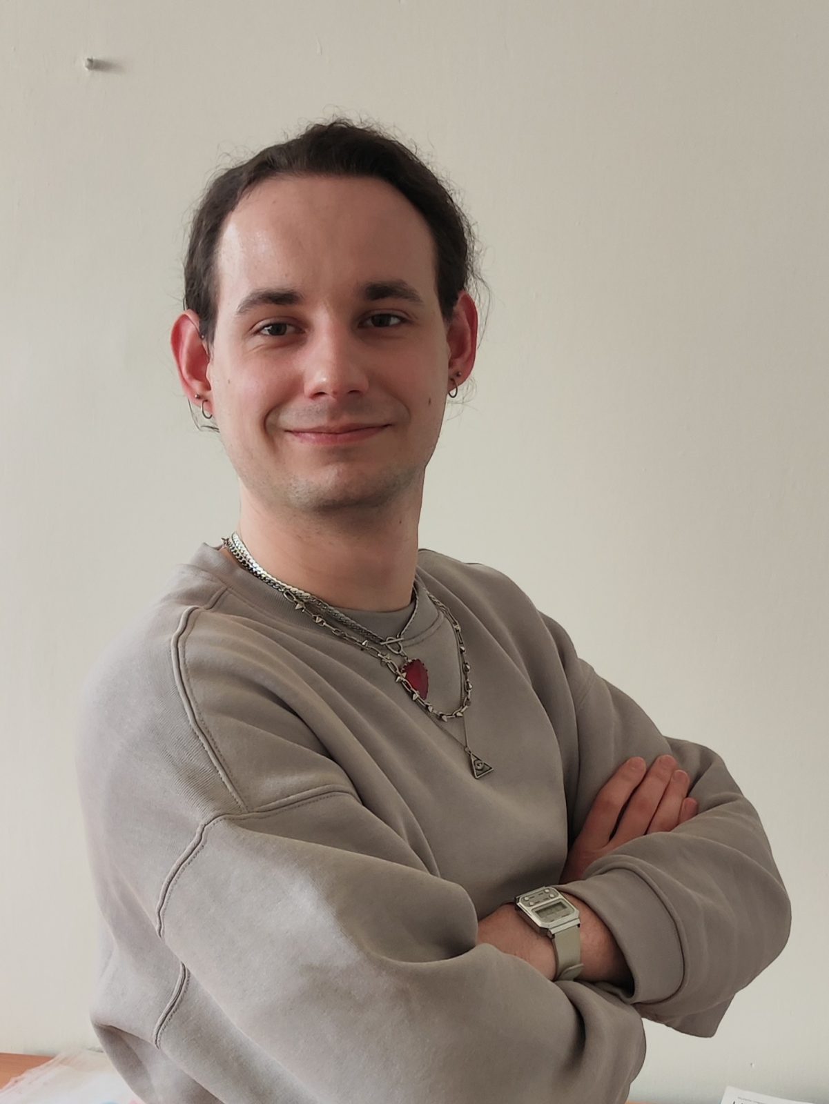 Uśmiechnięty młody mężczyzna, stoi na tle jasnej ściany z założonymi rękami. Ma na sobie luźną, beżową bluzę oraz kilka naszyjników, w tym jeden z czerwonym kamieniem.