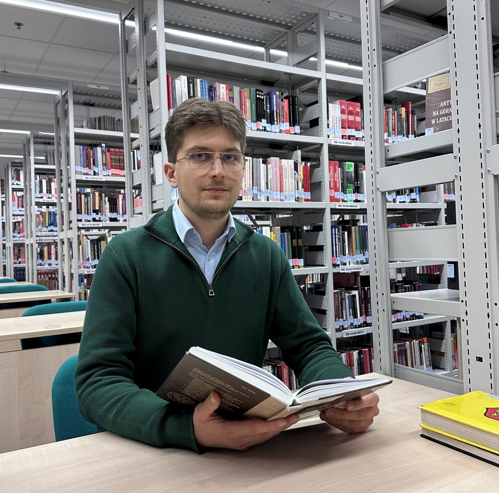 Młody mężczyzna siedzi przy biurku w bibliotece, trzymając otwartą książkę. Ma na sobie zielony sweter i jasną koszulę. W tle widać wysokie regały wypełnione książkami oraz rzędy stolików do pracy