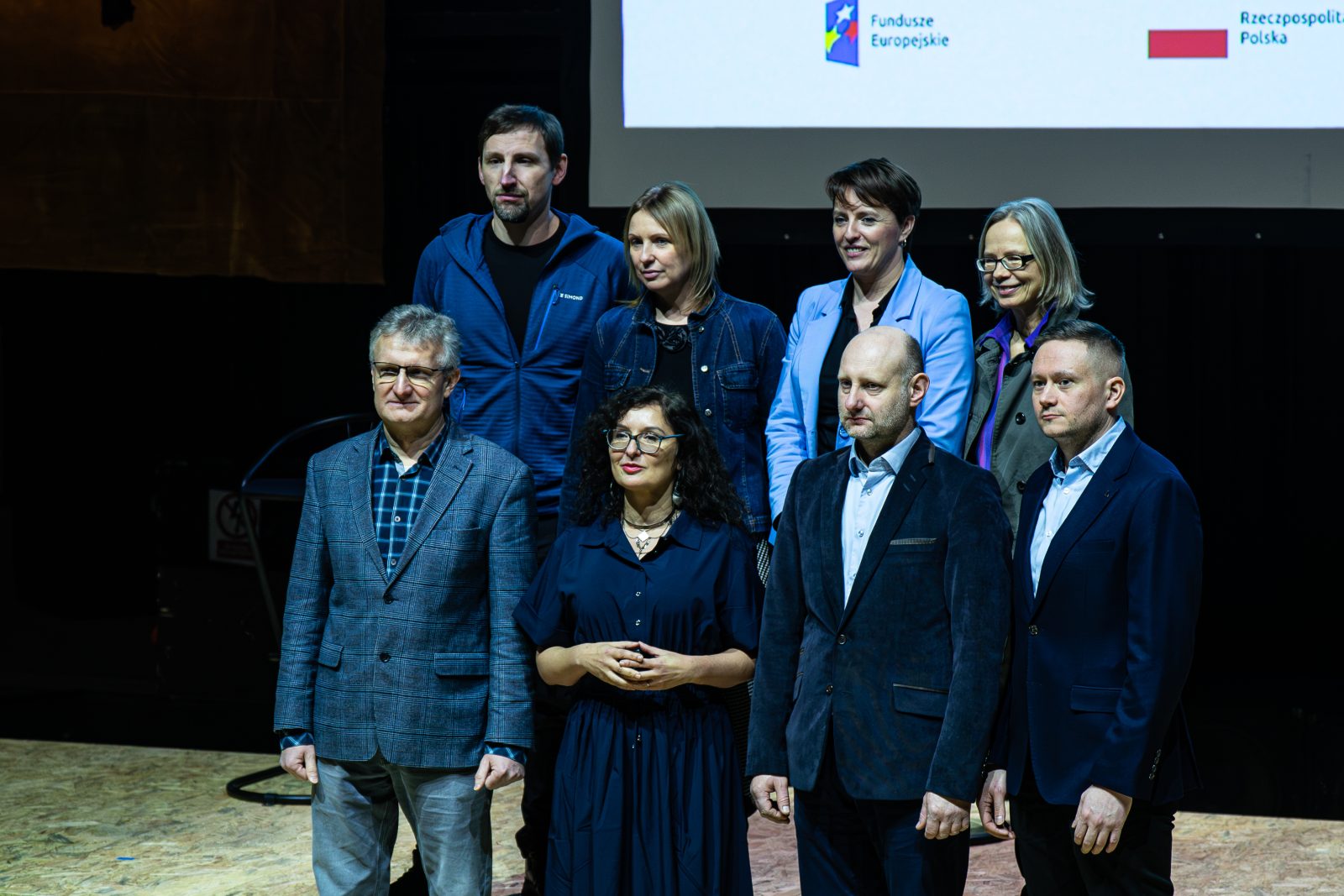 Zdjęcie przedstawia grupę osób stojących na scenie w dwóch rzędach podczas oficjalnego wydarzenia. Wszyscy są ubrani w eleganckie lub półformalne stroje, m.in. marynarki, koszule, sukienki i żakiety. Tło jest ciemne, a za grupą widoczny jest duży ekran projekcyjny z logotypami Funduszy Europejskich oraz Rzeczypospolitej Polskiej.