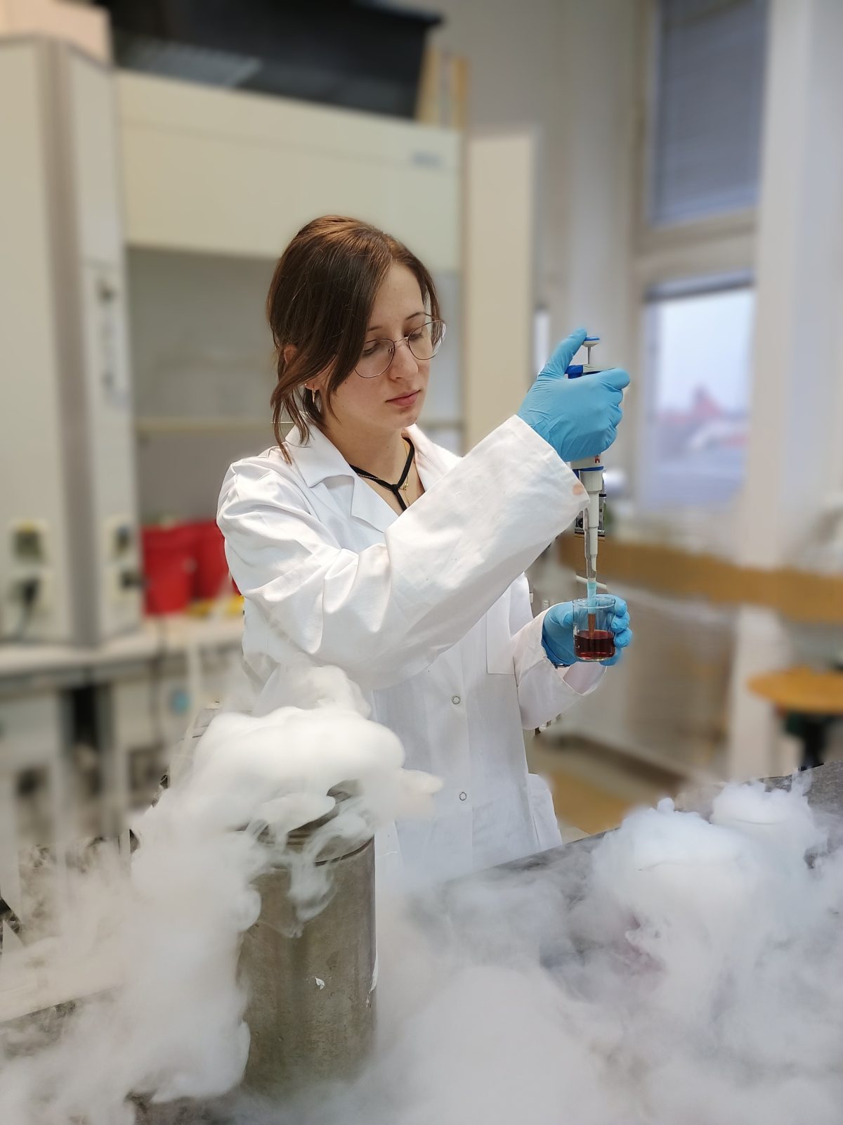 Młoda kobieta w białym fartuchu laboratoryjnym i niebieskich rękawiczkach wykonuje doświadczenie chemiczne przy stole w laboratorium. Trzyma w ręku pipetę, dozując ciecz do naczynia z czerwonym roztworem. Kadr wypełnia biały dym z innego naczynia laboratoryjnego.