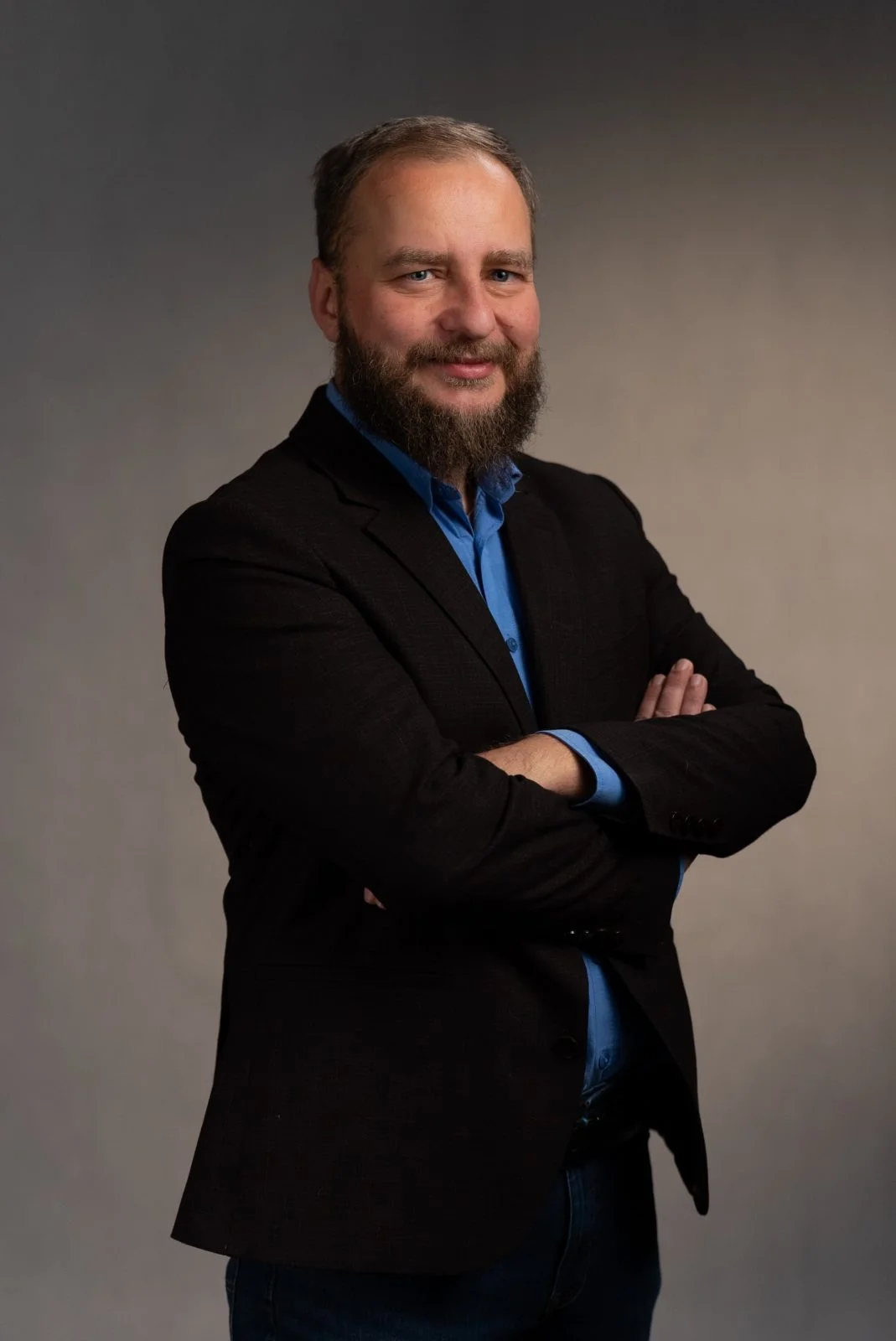 Mężczyzna w marynarce i koszuli stoi bokiem z założonymi rękami na neutralnym tle.