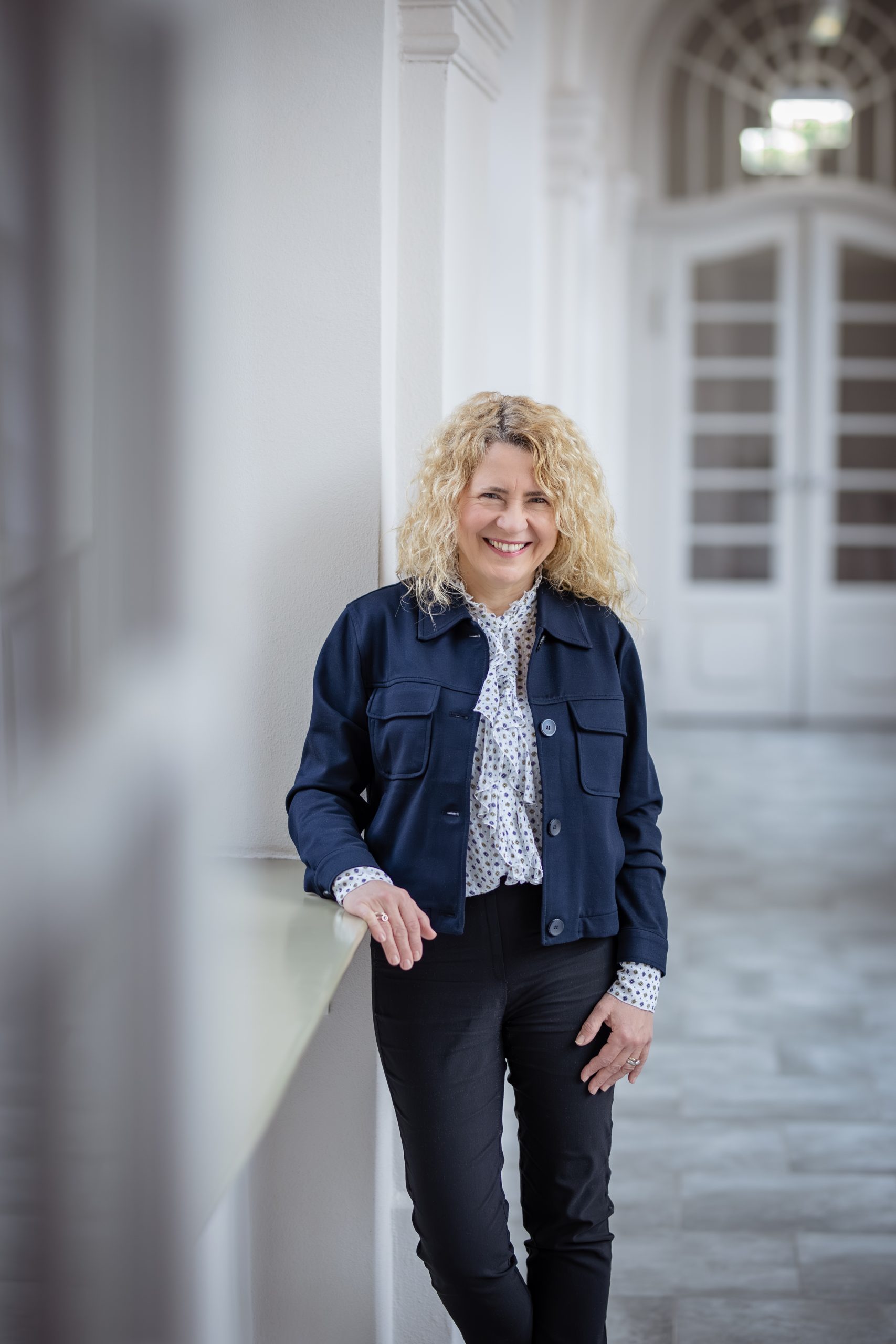 Uśmiechnięta kobieta z półdługimi, kręconymi, blond włosami, jasnej bluzce i ciemnogranatowej kurtce.