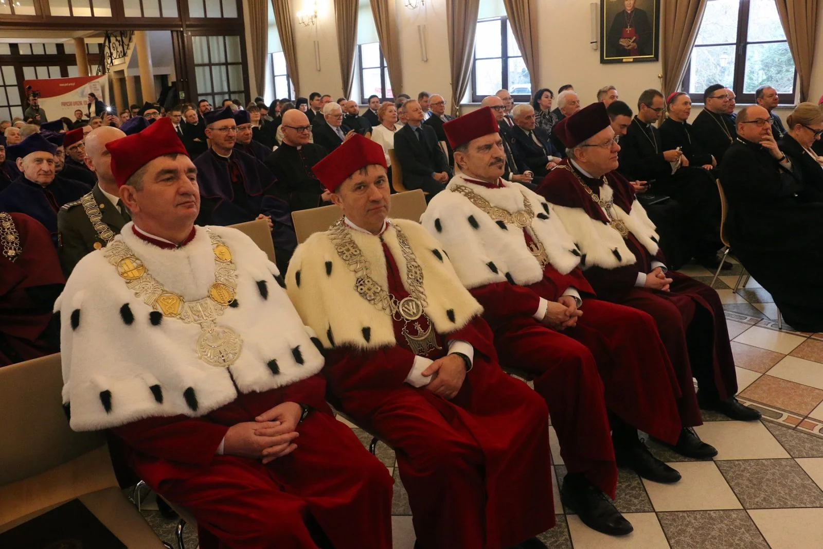 Duża sala, w której siedzi wiele osób. W pierwszym rzędzie siedzi kilku mężczyzn w uroczystych, czerwonych togach z białymi kołnierzami. Za nimi widać pełną salę uczestników uroczystości.