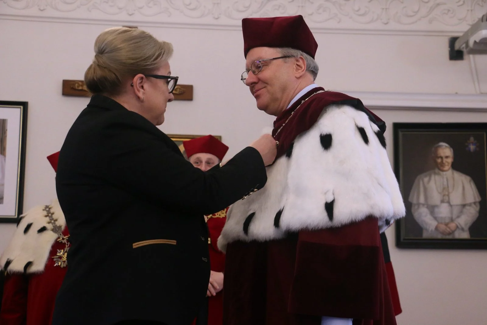 Kobieta przypina odznaczenie mężczyźnie w uroczystej czerwonej todze z białym kołnierzem. W tle widać inne osoby w podobnych strojach oraz portrety na ścianie.