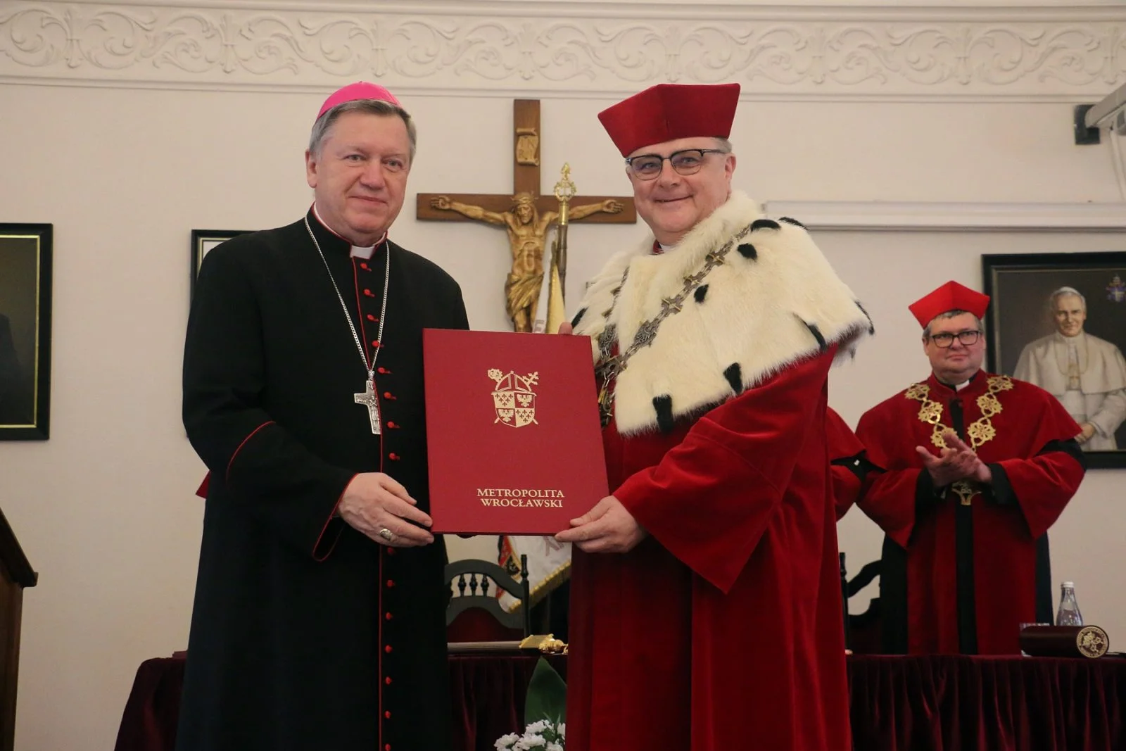 Dwaj mężczyźni w uroczystych strojach akademickich stoją obok siebie i trzymają duży czerwony dokument. W tle widać krucyfiks oraz innych uczestników ceremonii.