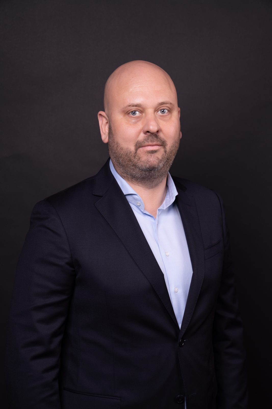 Mężczyzna z krótką brodą, łysy, ubrany w garnitur, z rękami splecionymi za plecami, stojący na czarnym tle.