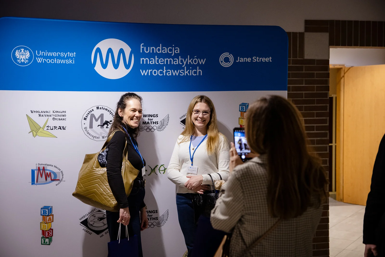 dwie osoby stoją na tle ścianki reklamowej Fundacji Matematyków Wrocławskich, trzecia osoba robi im zdjęcie telefonem komórkowym