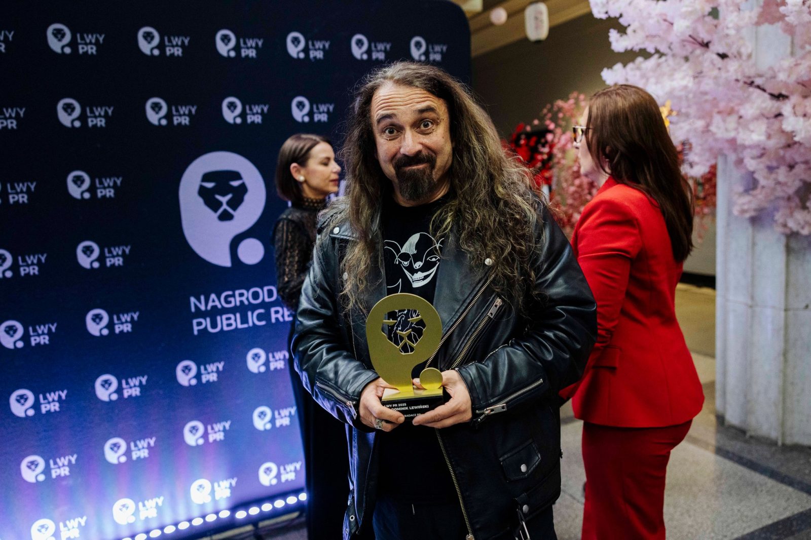 mężczyzna z długimi ciemnymi włosami i brodą, ubrany w skórzaną kurtkę, trzyma złotą statuetkę na tle ścianki promocyjnej z logotypem "LWY PR. Nagroda public relations"