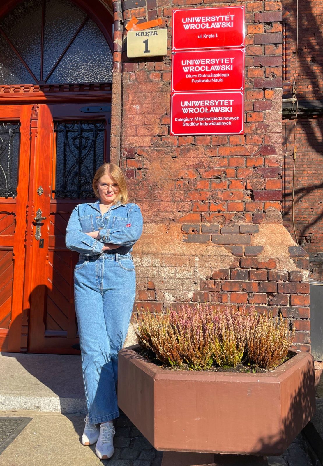 osoba w dżinsowej kurtce stojąca przed ceglaną ścianą z czerwoną tabliczką z napisem UNIWERSYTET WROCŁAWSKI Kolegium Międzydziedzinowych Studiów Indywidualnych