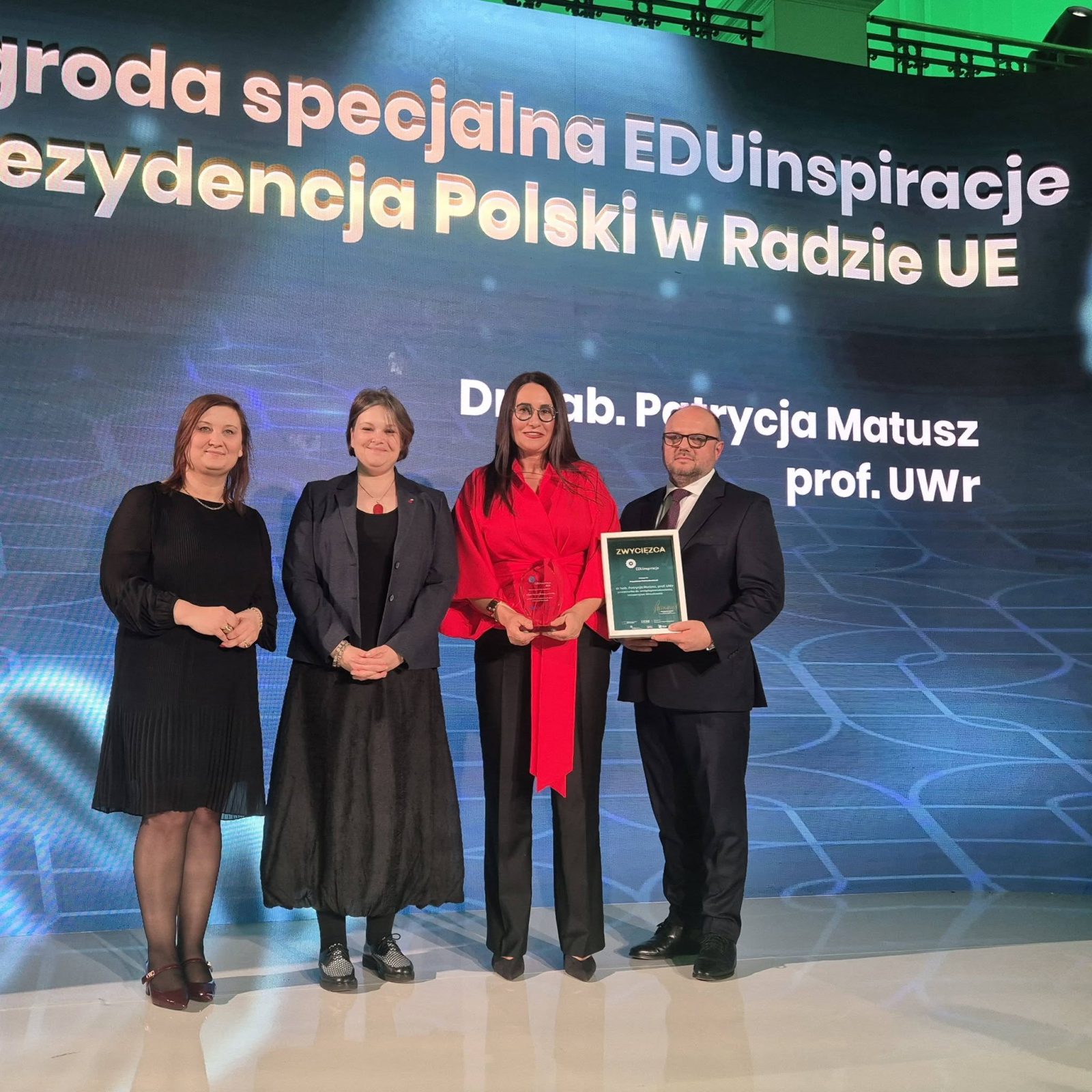 cztery osoby na scenie na tle ozdobnej ścianki, jedna z nich trzyma dyplom laureata konkursu
