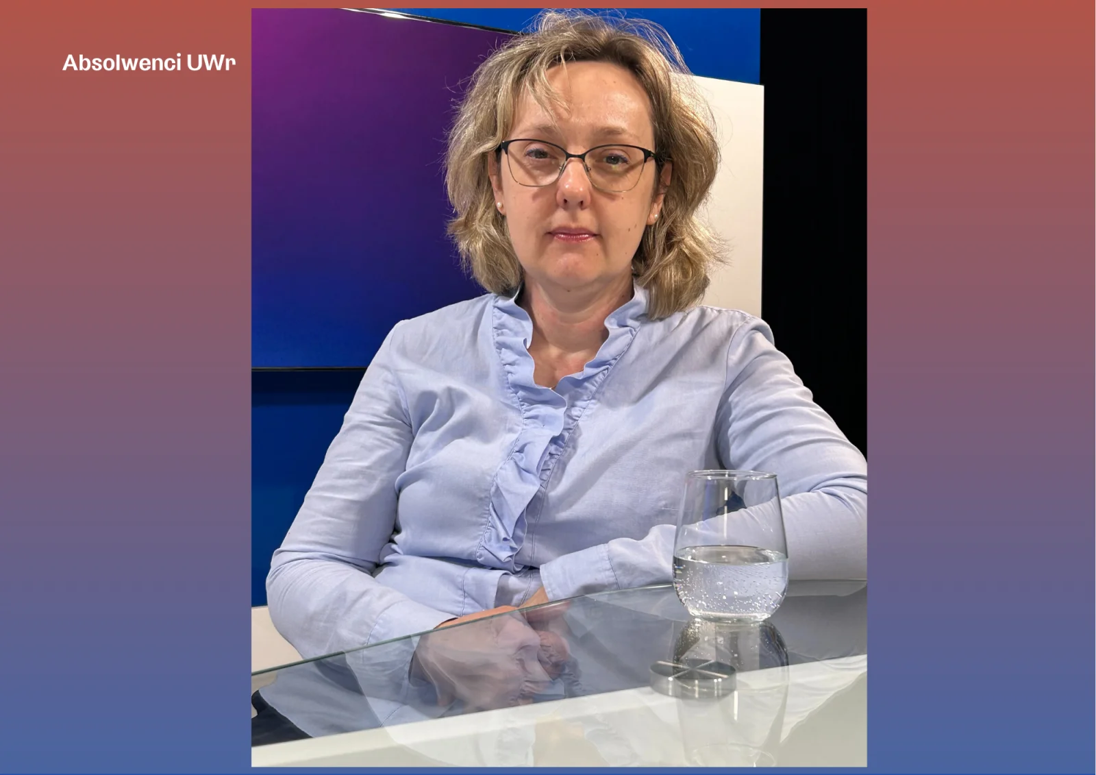 kobieta w koszuli i okularach siedząca przy stole, na którym stoi szklanka wody