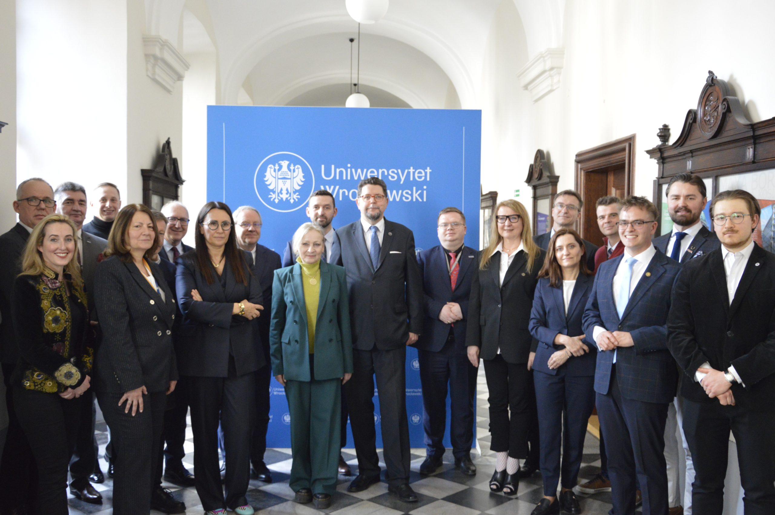 duża grupa elegancko ubranych osób na tle dużej niebieskiej ścianki z logotypem Uniwersytetu Wrocławskiego