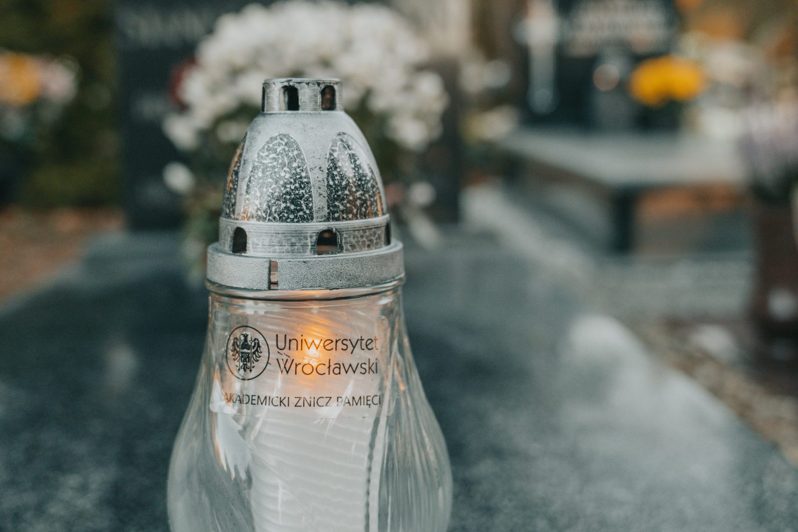 szklany znicz z szarą kopułą stojący na grobie. na zniczu znajduje się logo uniwersytetu wrocławskiego oraz napis akademicki znicz pamięci